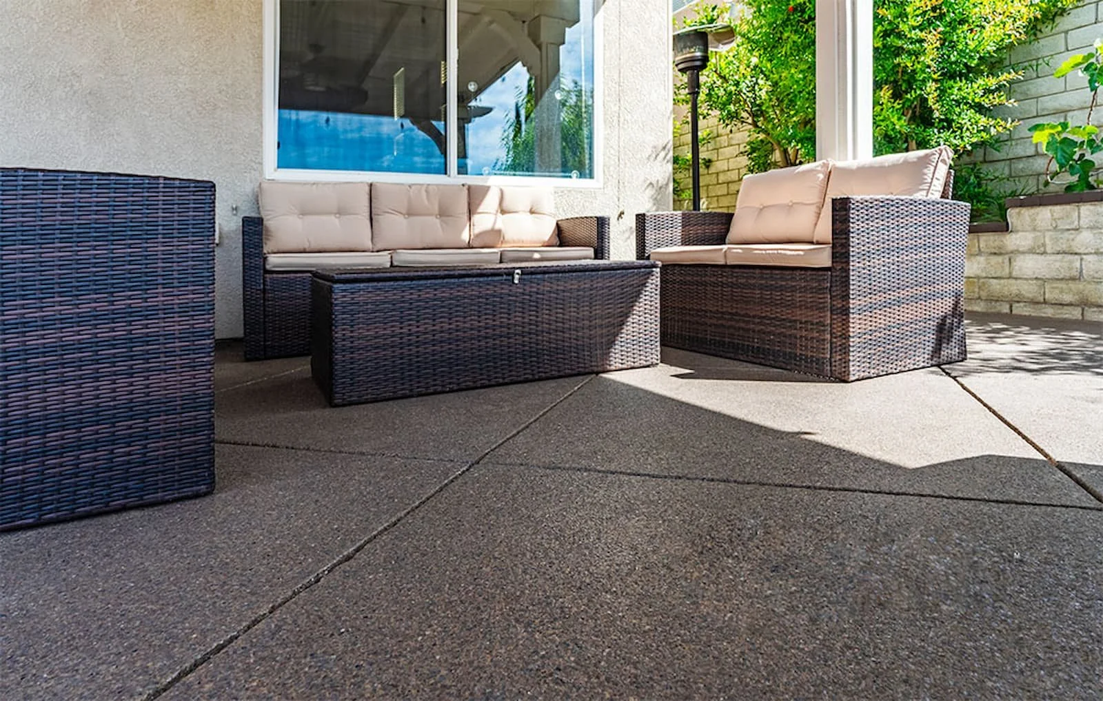 Outdoor patio with wicker furniture including a sofa, two armchairs, and an ottoman, with a beige cushion on the furniture and green plants along a brick wall in the background.