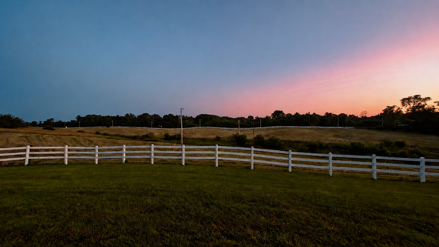 Sunset over a rural landscape with rolling hills, a white fence, and trees, with a clear sky transitioning from blue to pink and orange.