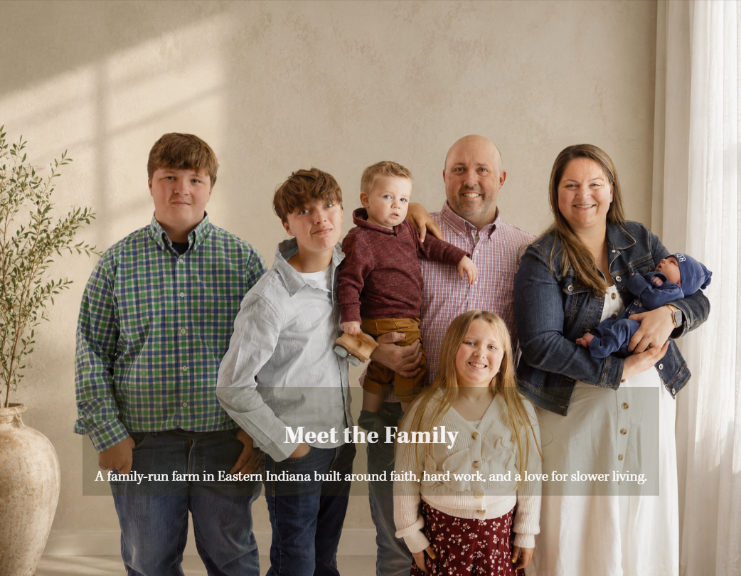 A family of seven standing together in a room with beige walls and white curtains, including two young children, a couple, and one baby being held by the woman.