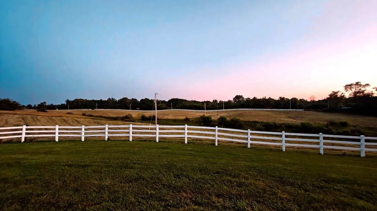 Open landscape with grassy foreground, white fence, rolling hills, trees, and a sky transitioning from blue to pink at sunset.
