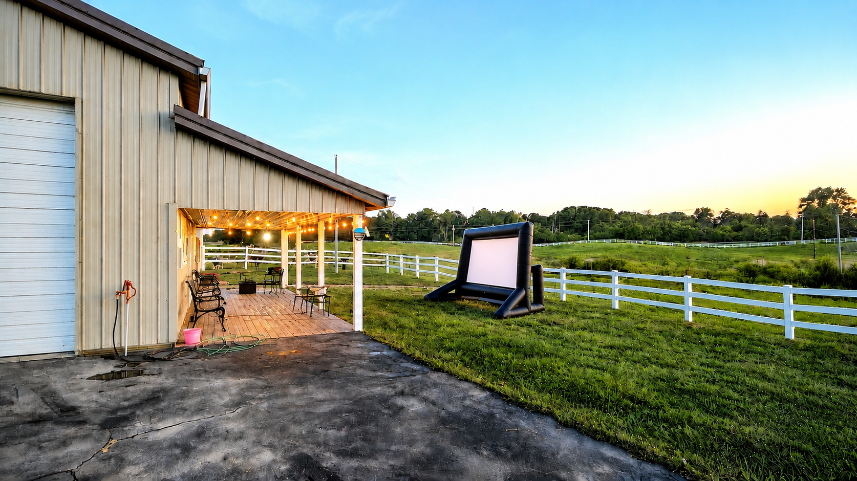 A backyard patio with string lights, chairs, and a grassy field with a white fence in the background during sunset.