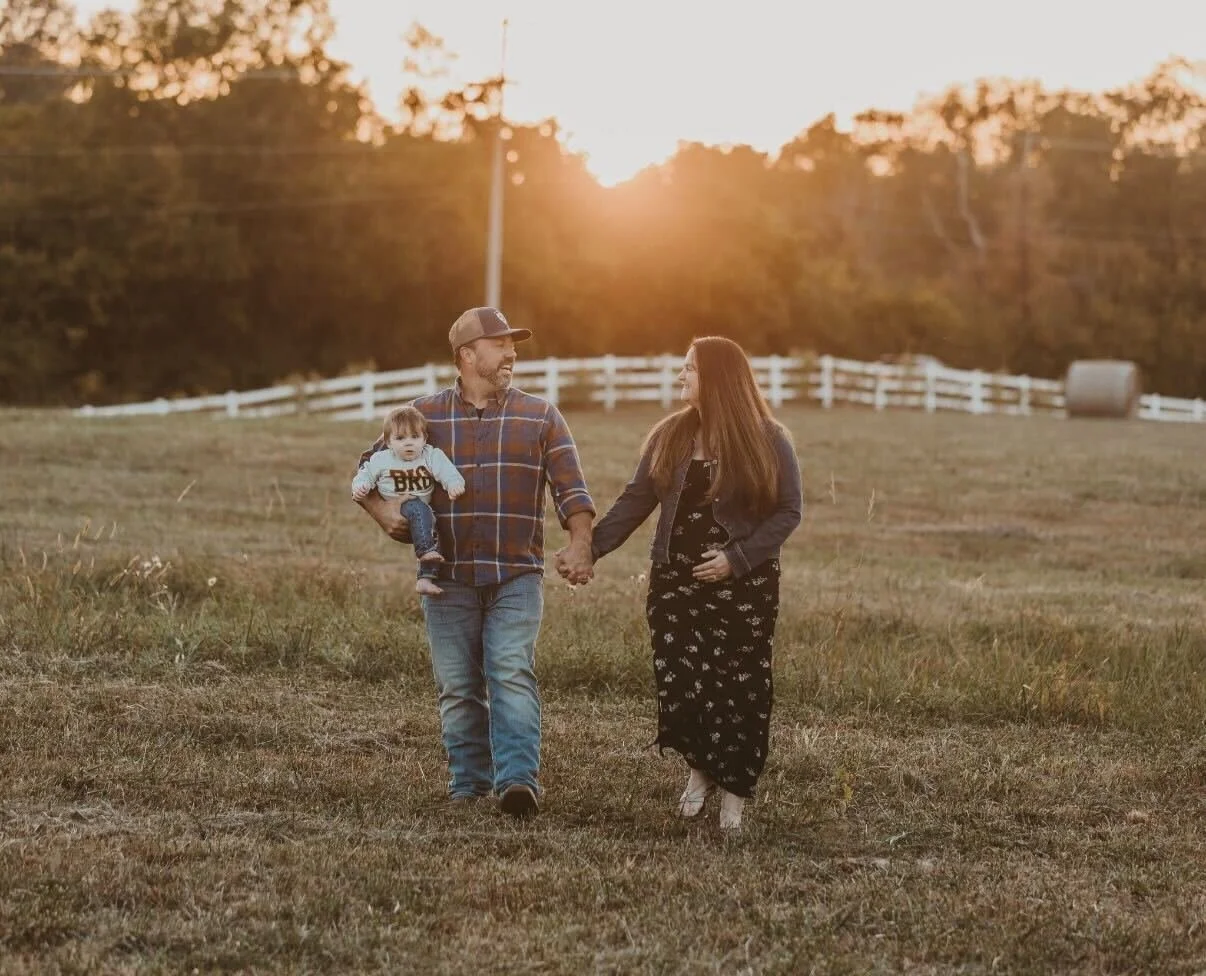 A family walking hand in hand in a grassy field during sunset, with a white fence and trees in the background.