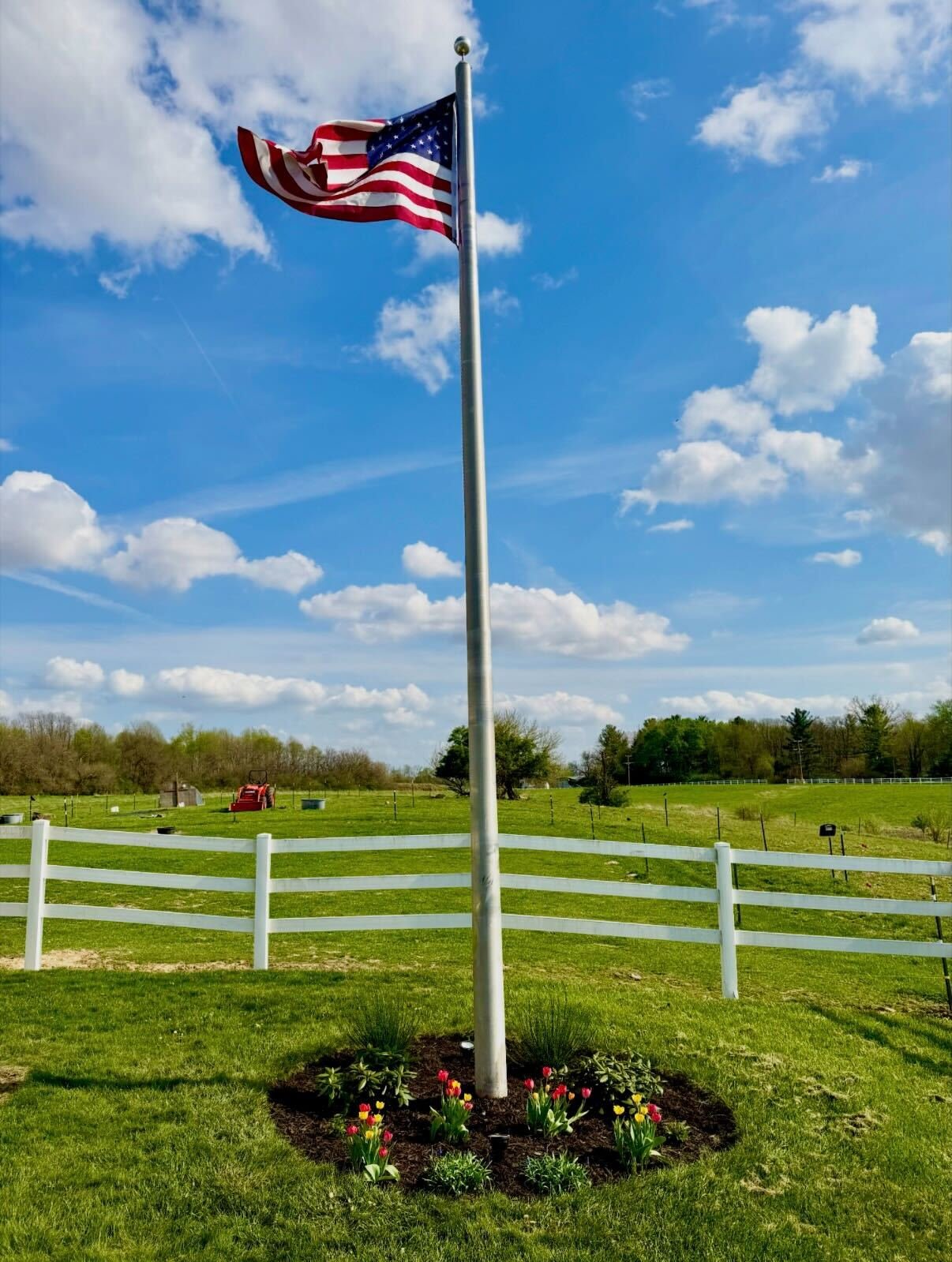 American flag on a flagpole in a grassy yard with a white fence, under a blue sky with scattered clouds.