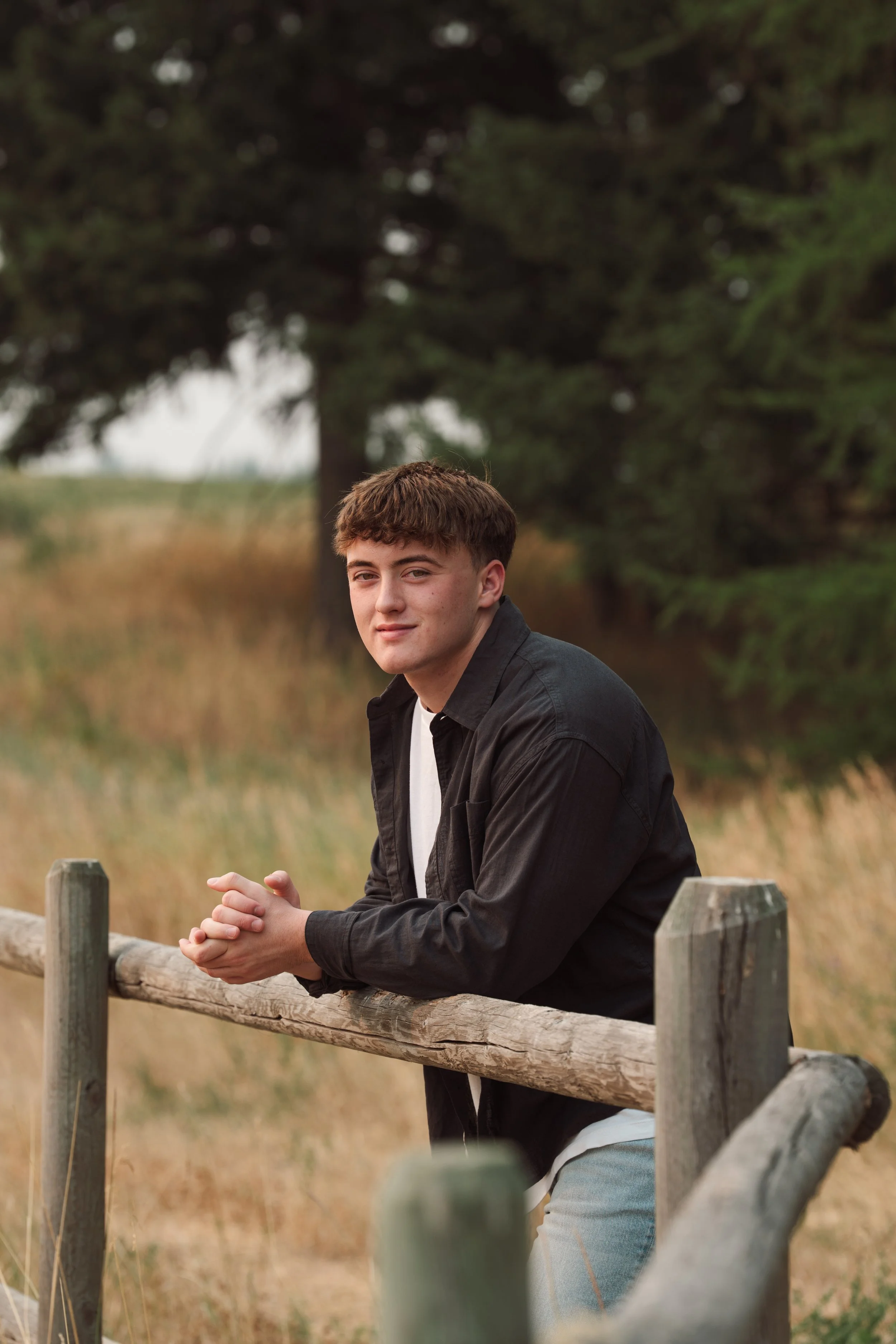 A young man with brown hair leaning on a wooden fence outdoors, with trees and grass in the background.