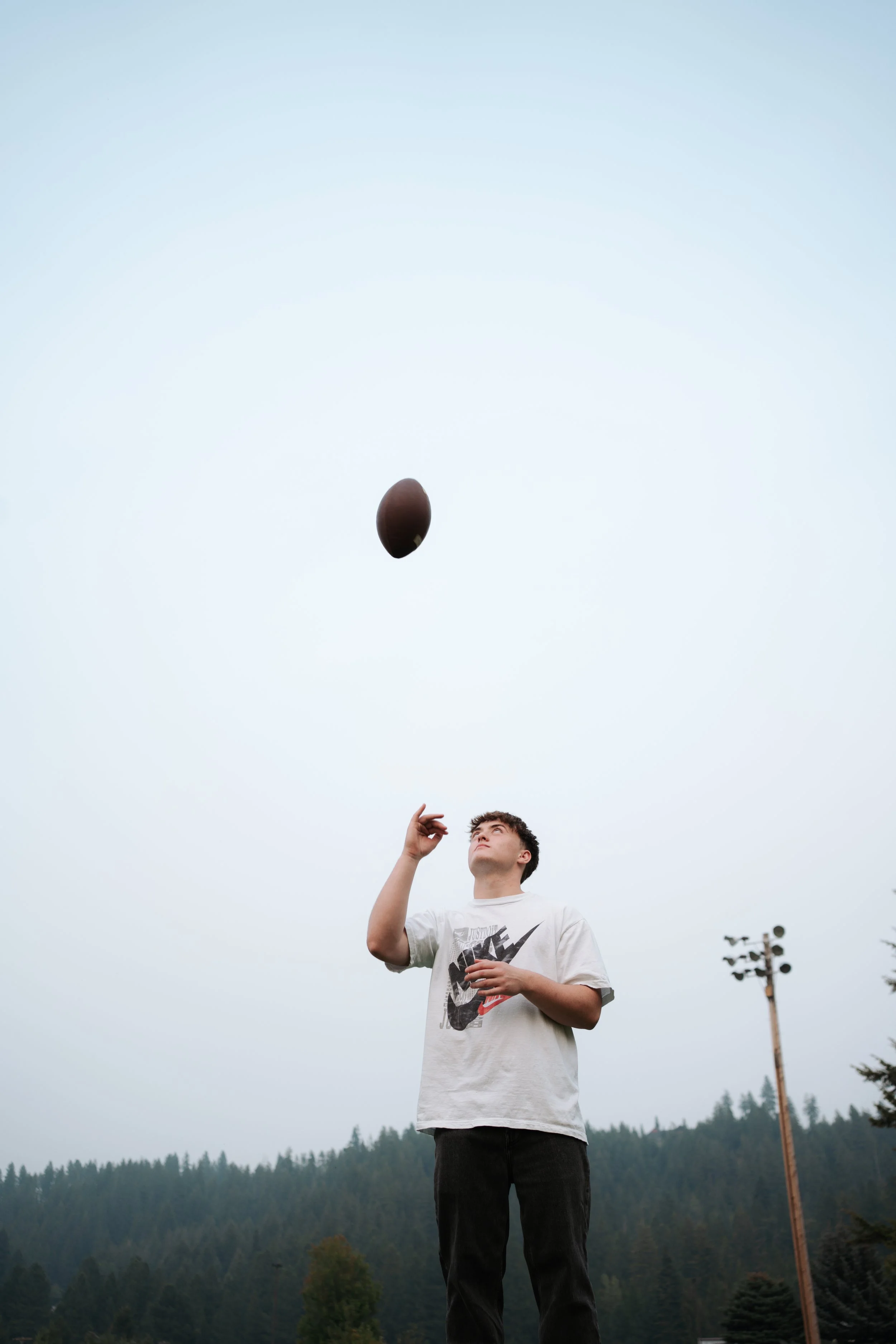 A young person wearing a Nike t-shirt is outdoors playing football, tossing the ball into the air against a cloudy sky with trees and a light pole in the background.