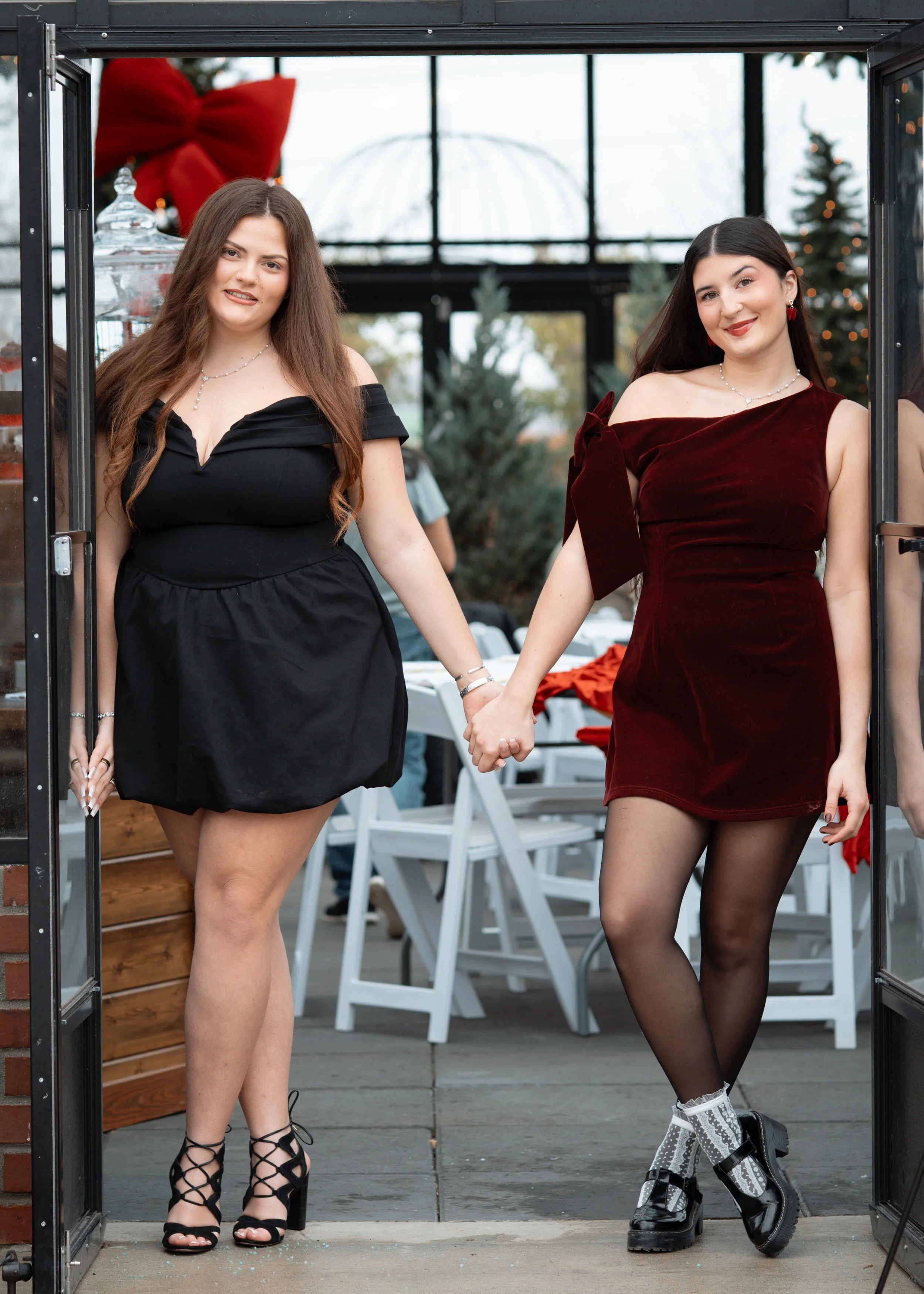 Two women holding hands and standing in a doorway at a festive celebration, with Christmas decorations visible in the background.