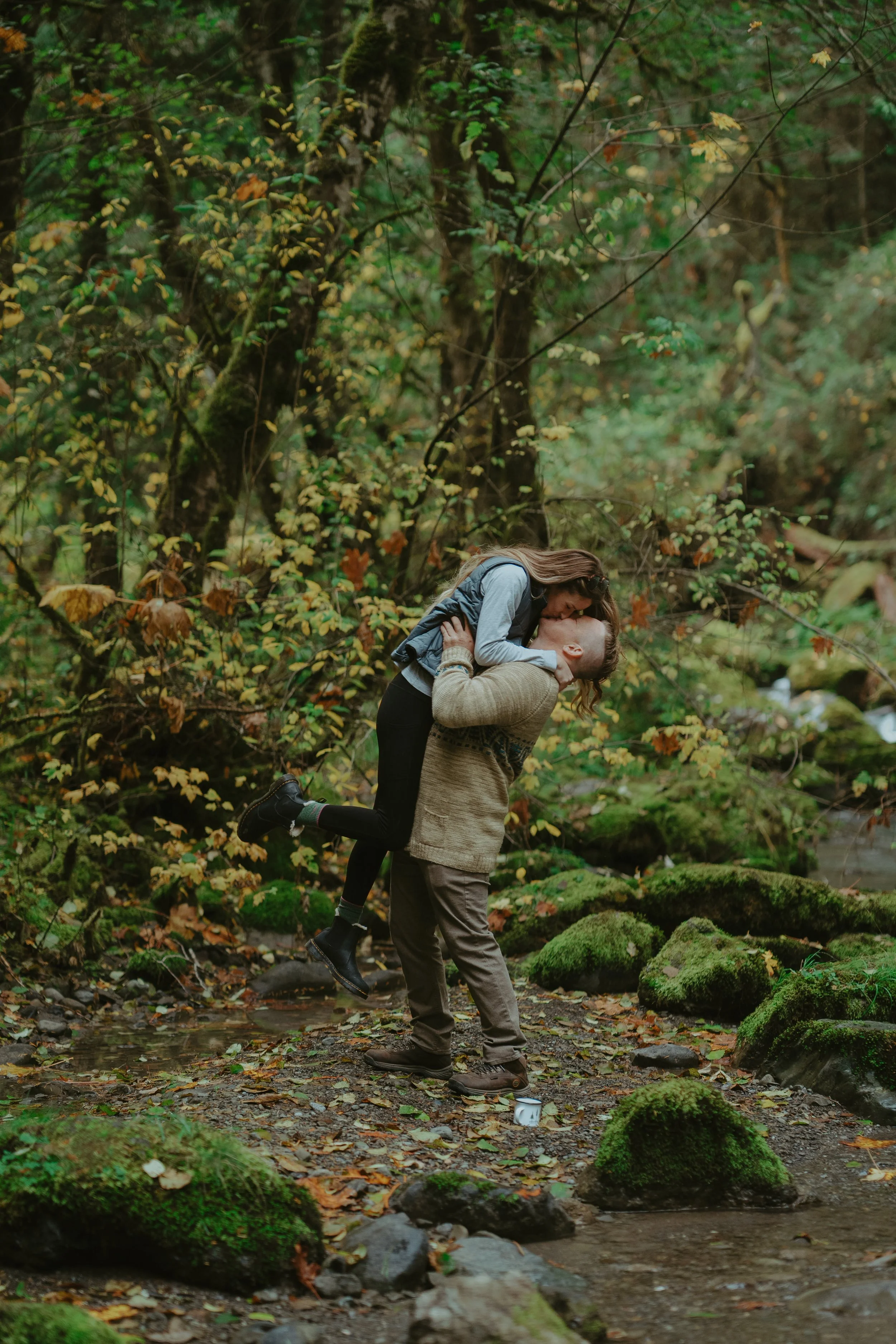 A man is holding a woman in his arms in a forest, and they are kissing while standing on a rocky, leaf-covered ground by a stream.