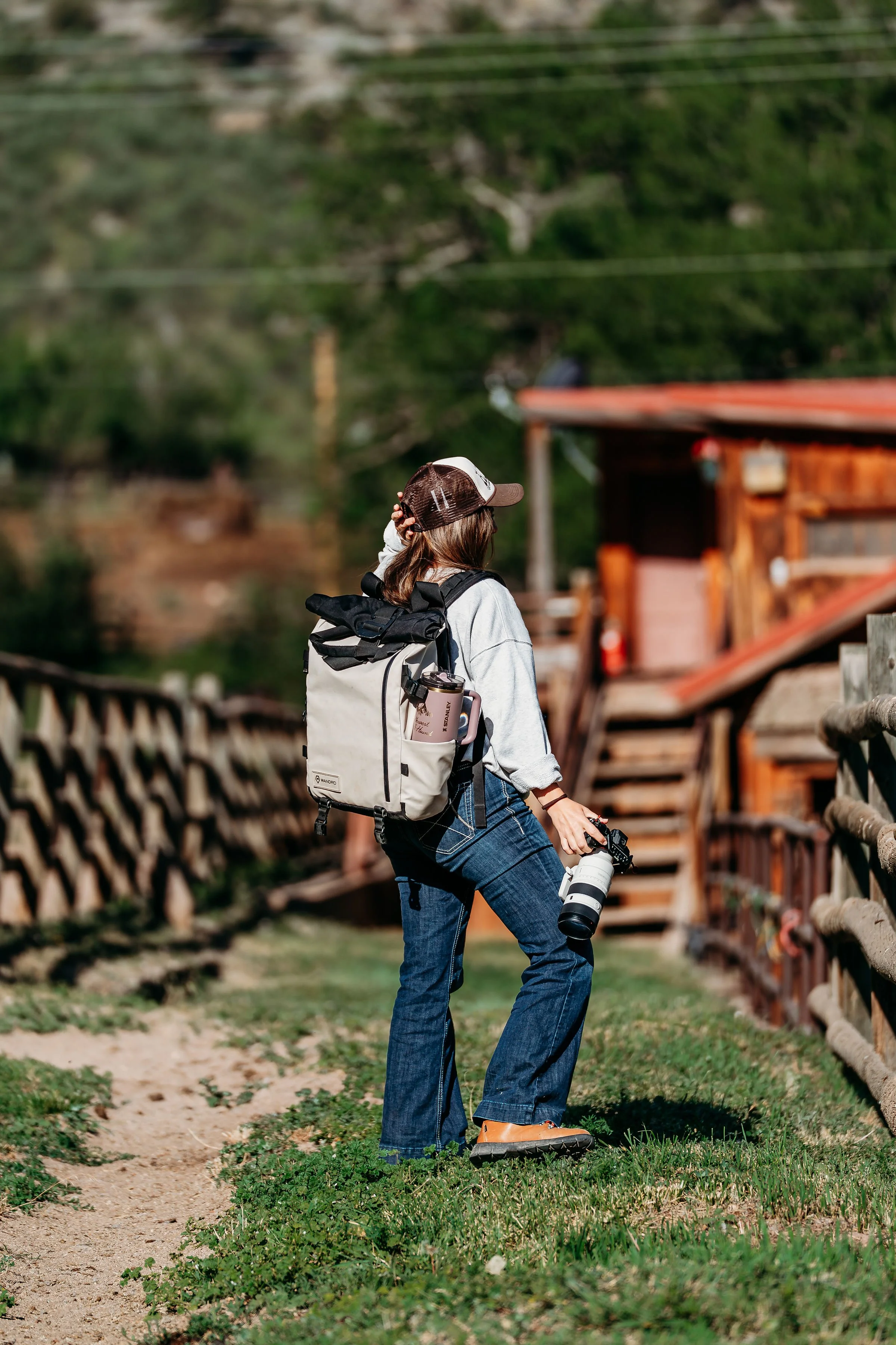 A woman in casual clothing, wearing a baseball cap, backpack, and holding a camera with a long lens, stands on a dirt path, looking into the distance. She is surrounded by greenery and wooden structures, with power lines on a hillside in the background.