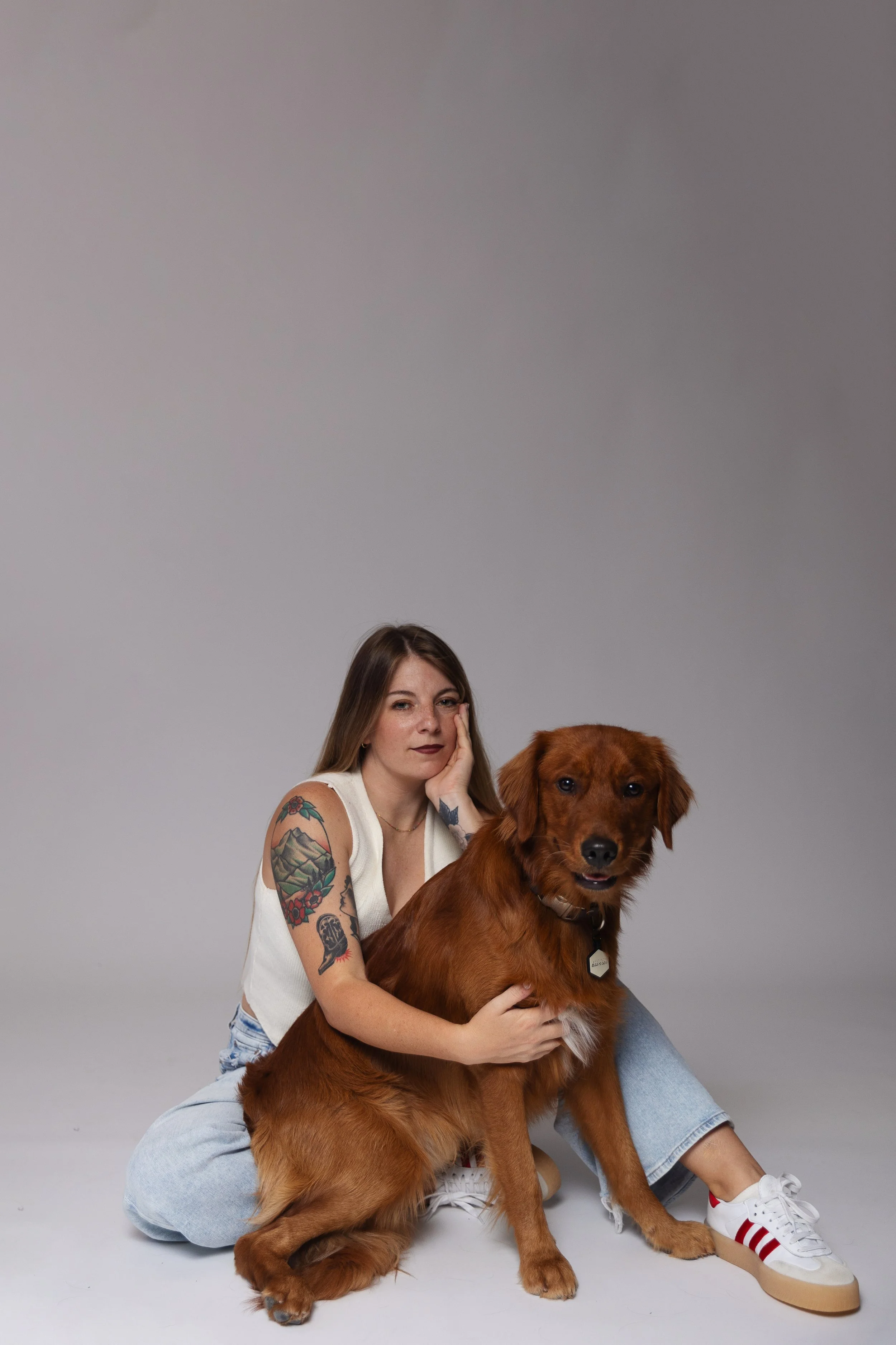 A young woman with long brown hair and tattoos on her arm, sitting in a relaxed pose, holding a large brown dog with long fur. The woman is wearing a white sleeveless top and light blue jeans. The dog appears happy with a slightly open mouth, and grey eyes, wearing a collar with a tag. The background is plain grey, and the woman is wearing white sneakers with red stripes.