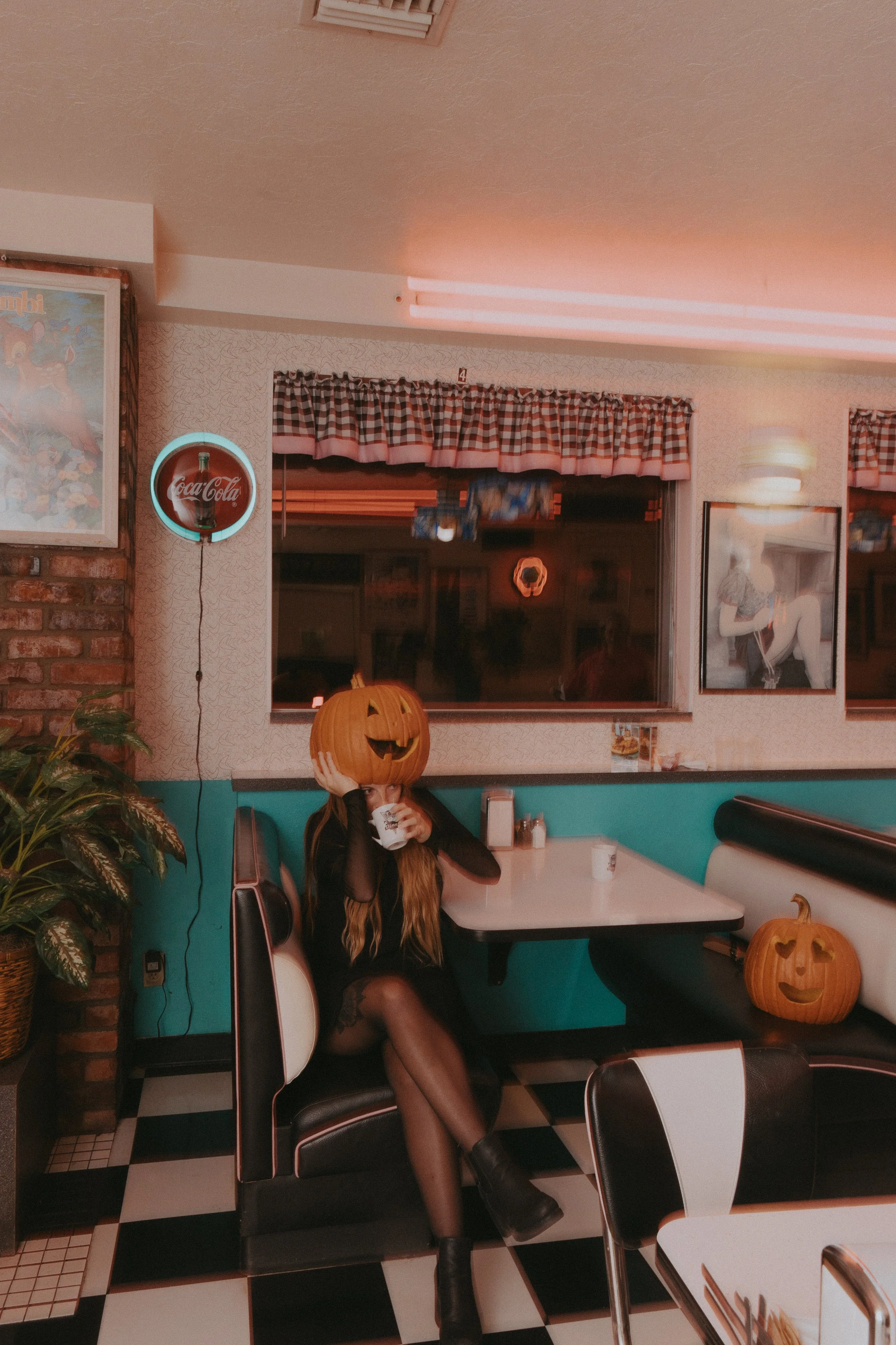 A woman with long blonde hair sitting in a retro-style diner booth, wearing a black dress and fishnet stockings, holding a paper cup with her face obscured by a carved pumpkin Halloween decoration. The diner has black and white checkered flooring, teal and white walls, checkered curtains, and Halloween decorations including a pumpkin on the floor.
