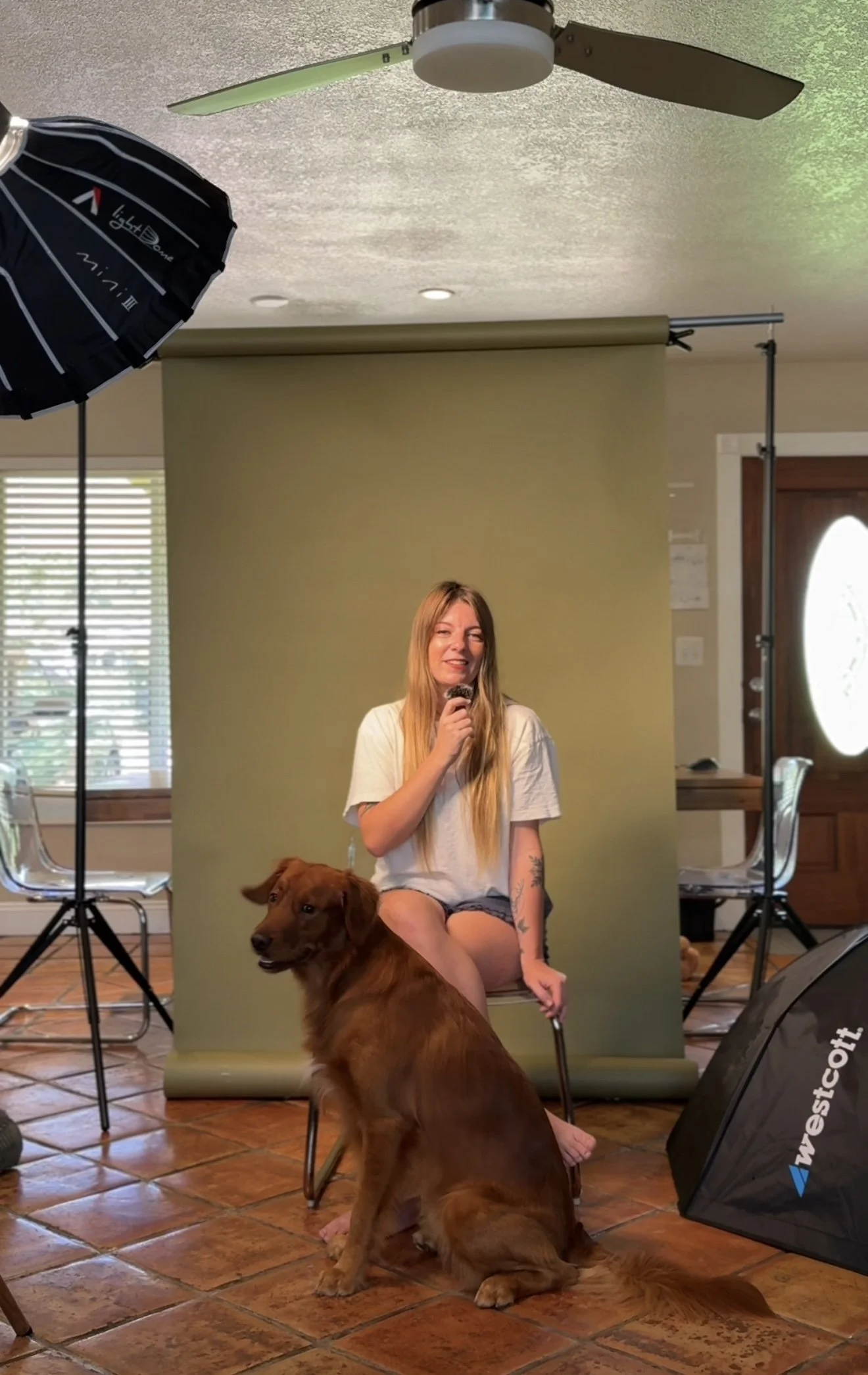 A woman with long blonde hair sitting on a chair with a brown dog in front of a green backdrop. The woman is holding a microphone and smiling, wearing a white t-shirt and shorts. The setting appears to be a home photography or studio setup with lighting equipment and a window with blinds.