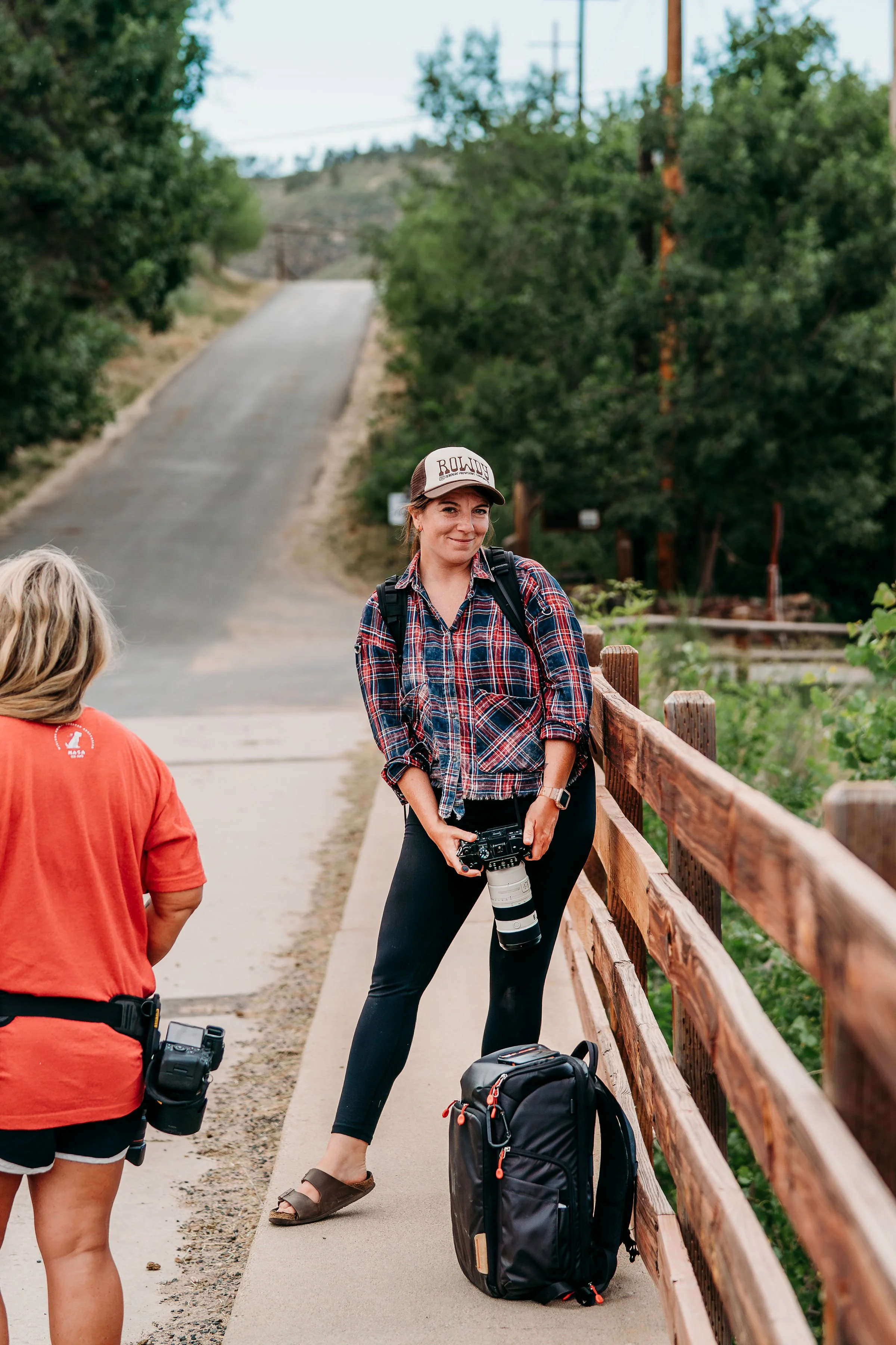A woman with a backpack and a camera standing next to a wooden fence on a hiking trail, smiling, with another person partially visible in the foreground.