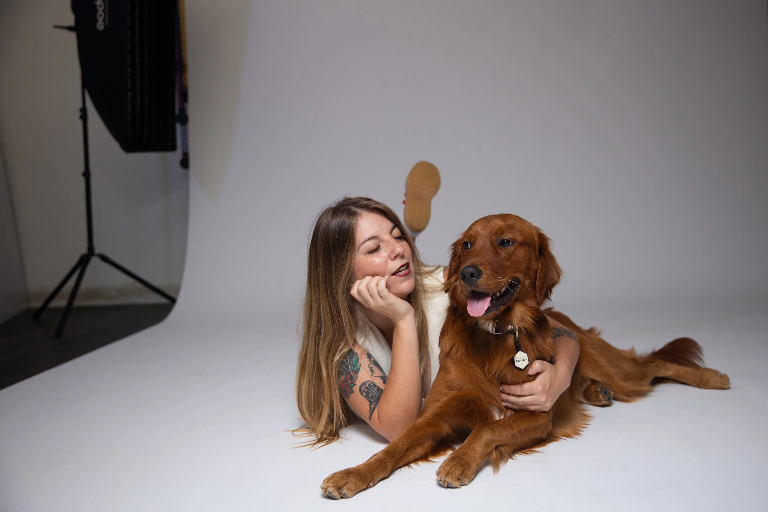 A woman with long hair and tattoos is lying on a white floor, holding a large golden retriever dog with a tag on its collar. The woman and dog are looking content, with the dog panting. Studio lighting equipment is visible in the background, indicating a photo shoot setting.
