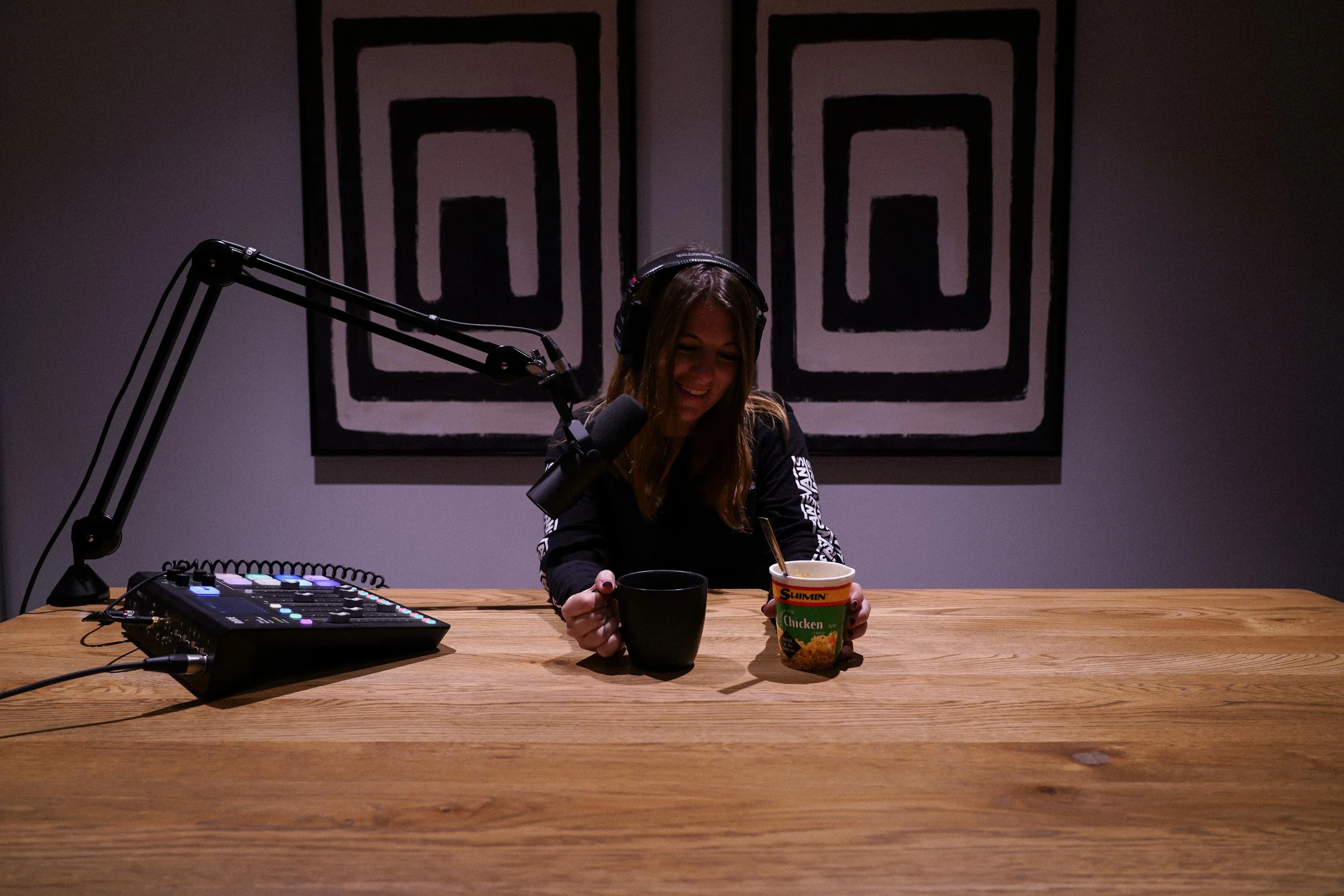 A woman with headphones sitting at a wooden table with a microphone, a music controller, and two cups, one black and one with chicken noodle soup. She is smiling and looking down.