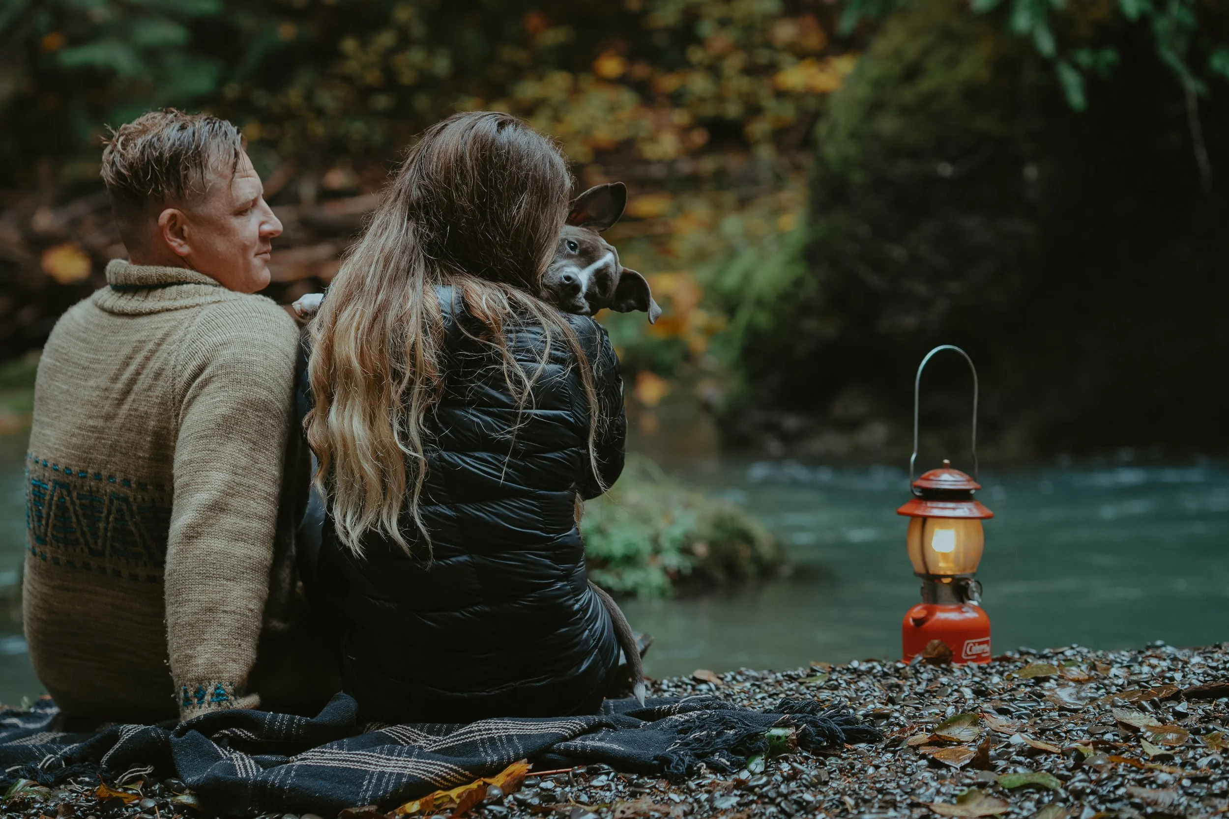 A man and woman sit on a blanket by a river surrounded by autumn trees, with a lantern nearby, as they enjoy the scenic outdoor setting with a dog.