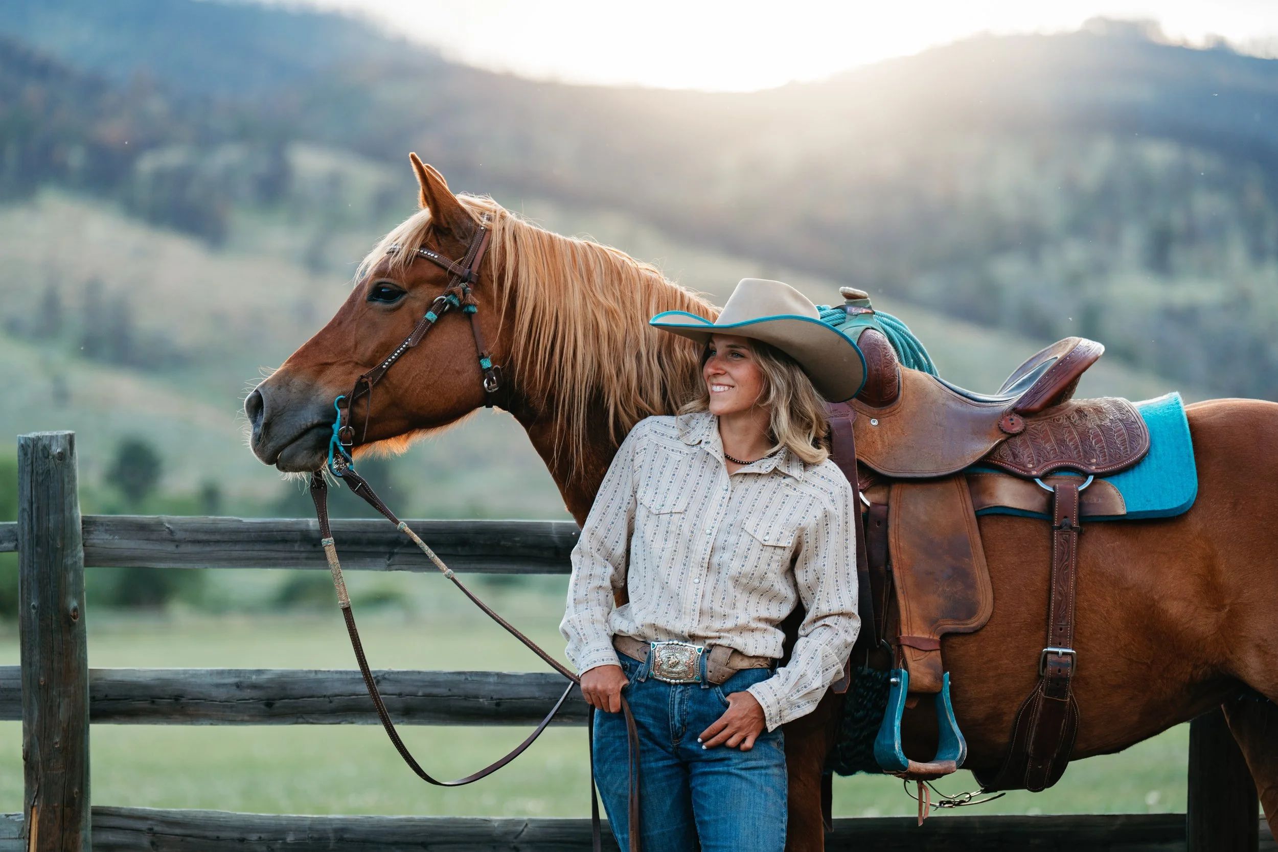 A woman in a cowboy hat smiling while standing next to a horse. The horse is saddled and bridled, with a blue blanket underneath the saddle, and is looking to the side. The woman is wearing a light-colored button-up shirt and jeans, standing next to a wooden fence with rolling hills in the background.