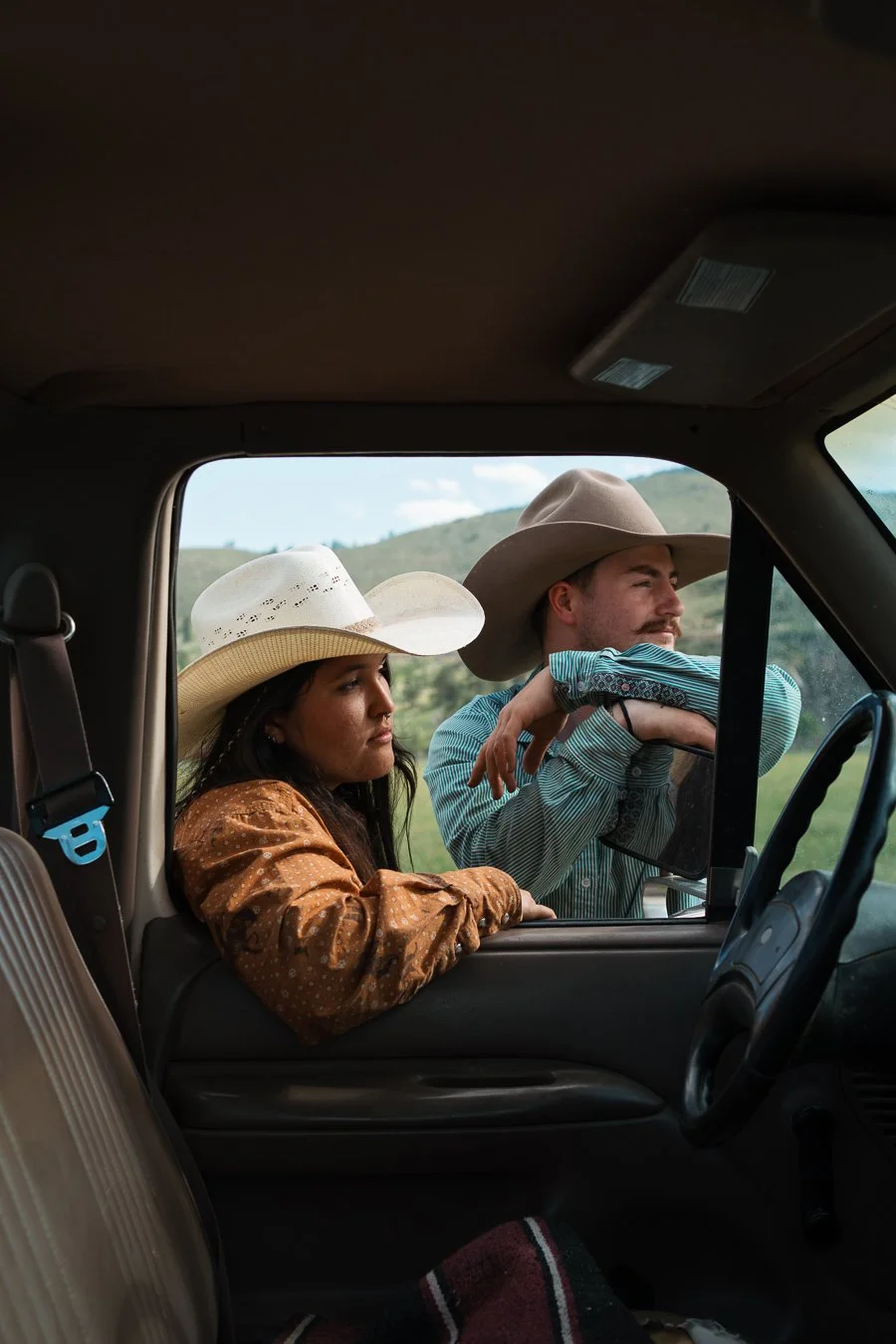 A man and woman wearing cowboy hats, sitting in a vehicle, looking out over a rural landscape with green hills and partly cloudy sky.