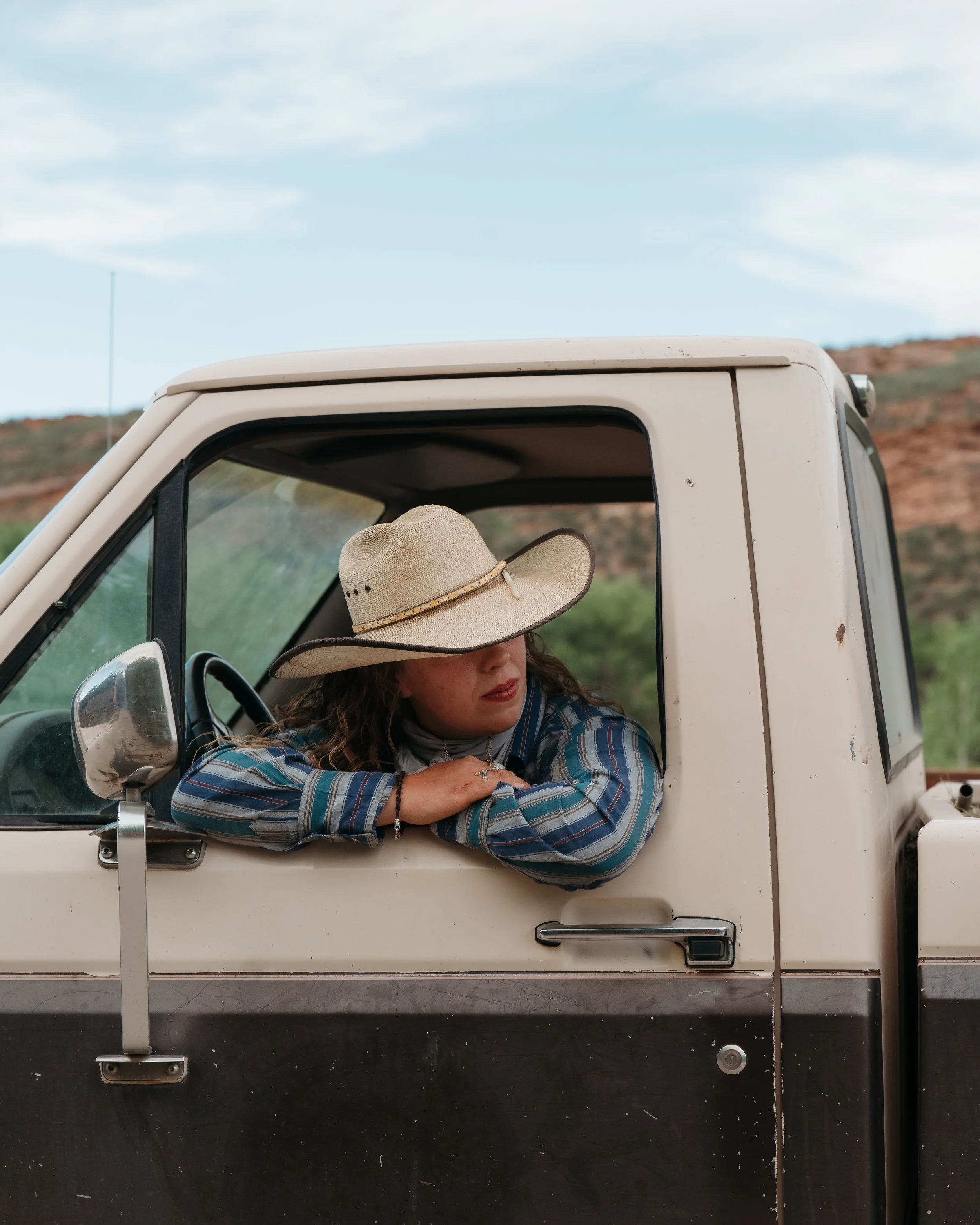 A woman with curly hair wearing a big straw hat and a plaid shirt, sitting in the driver's seat of a white pickup truck, with her arm resting on the window frame.