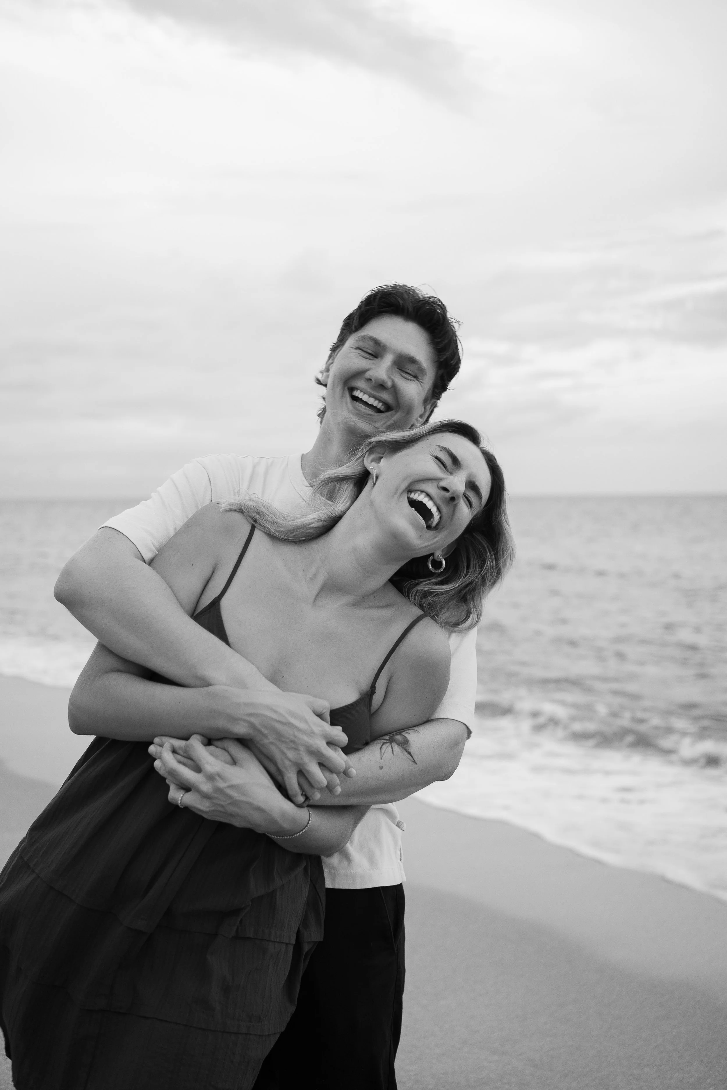 A smiling couple with closed eyes hugging on a beach, ocean waves and cloudy sky in the background, in black and white.