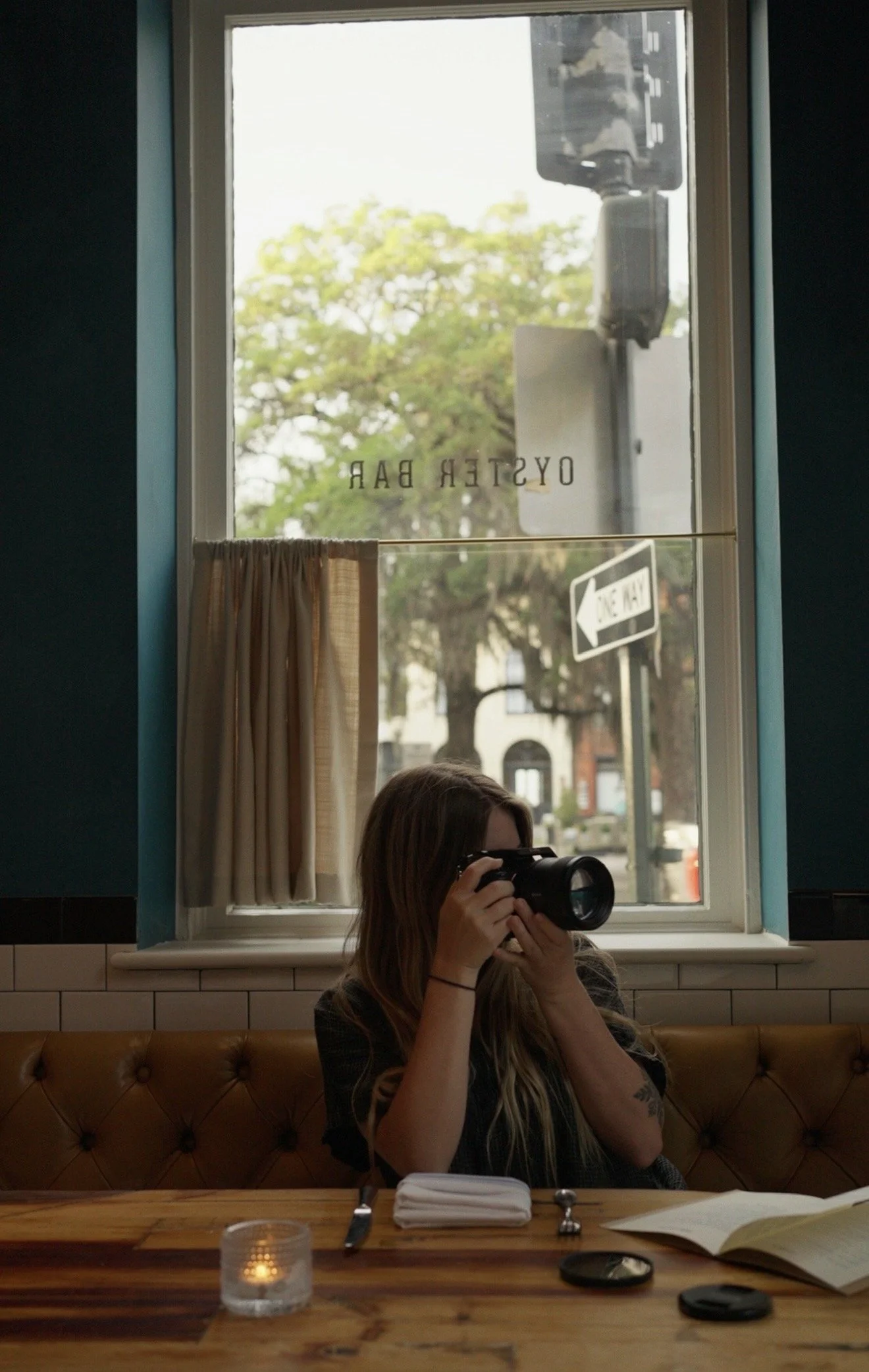 A woman with long hair and glasses is sitting at a restaurant table, holding a camera in front of her face. The window behind her shows a view of trees, a street sign, and a traffic light outside.