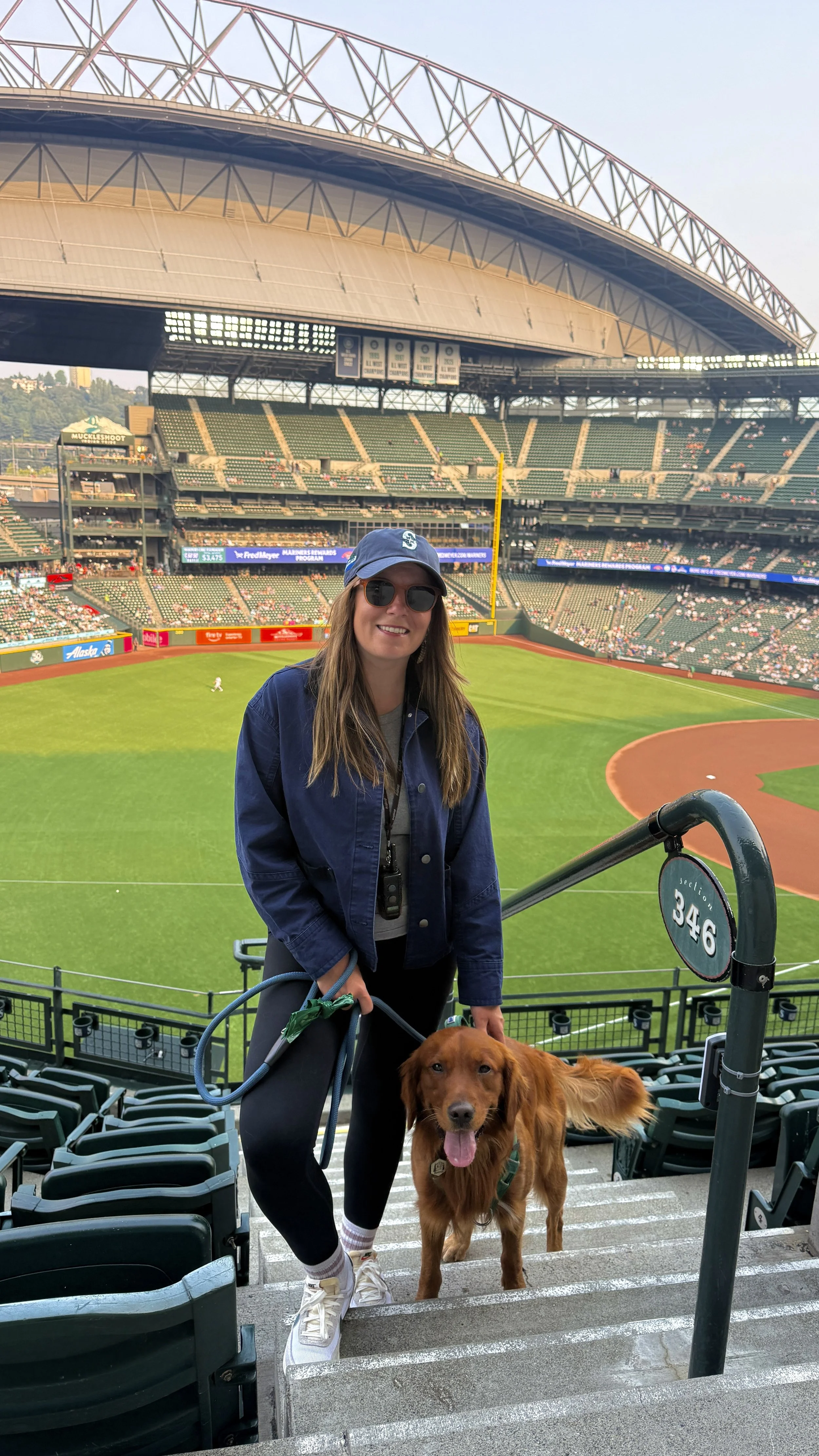 A woman in sunglasses and a baseball cap smiles at the camera while standing with a golden retriever dog on a staircase at a baseball stadium. The stadium is mostly empty with green seats and a view of the field in the background.