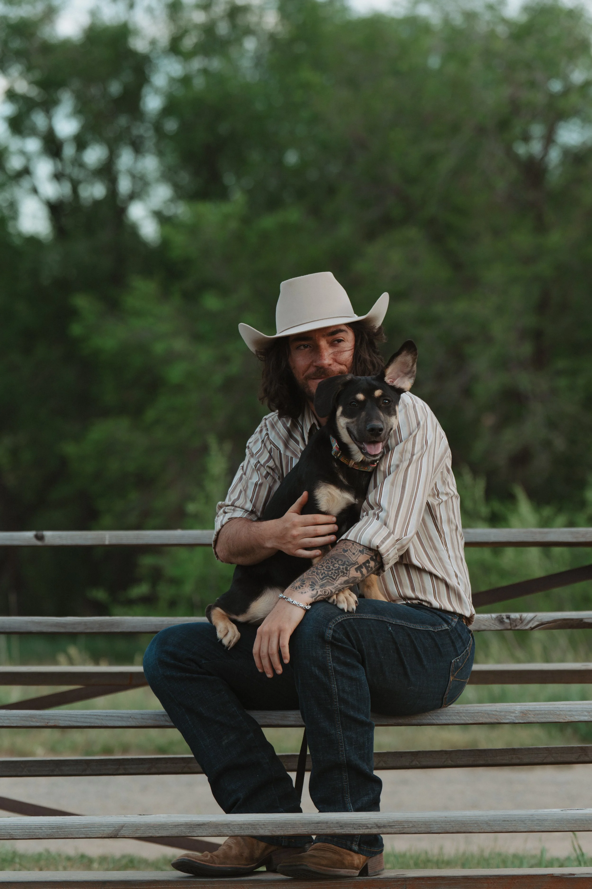 A man wearing a cowboy hat and striped shirt sitting on a wooden bench outdoors, holding a black and tan dog with one ear standing up, with greenery in the background.