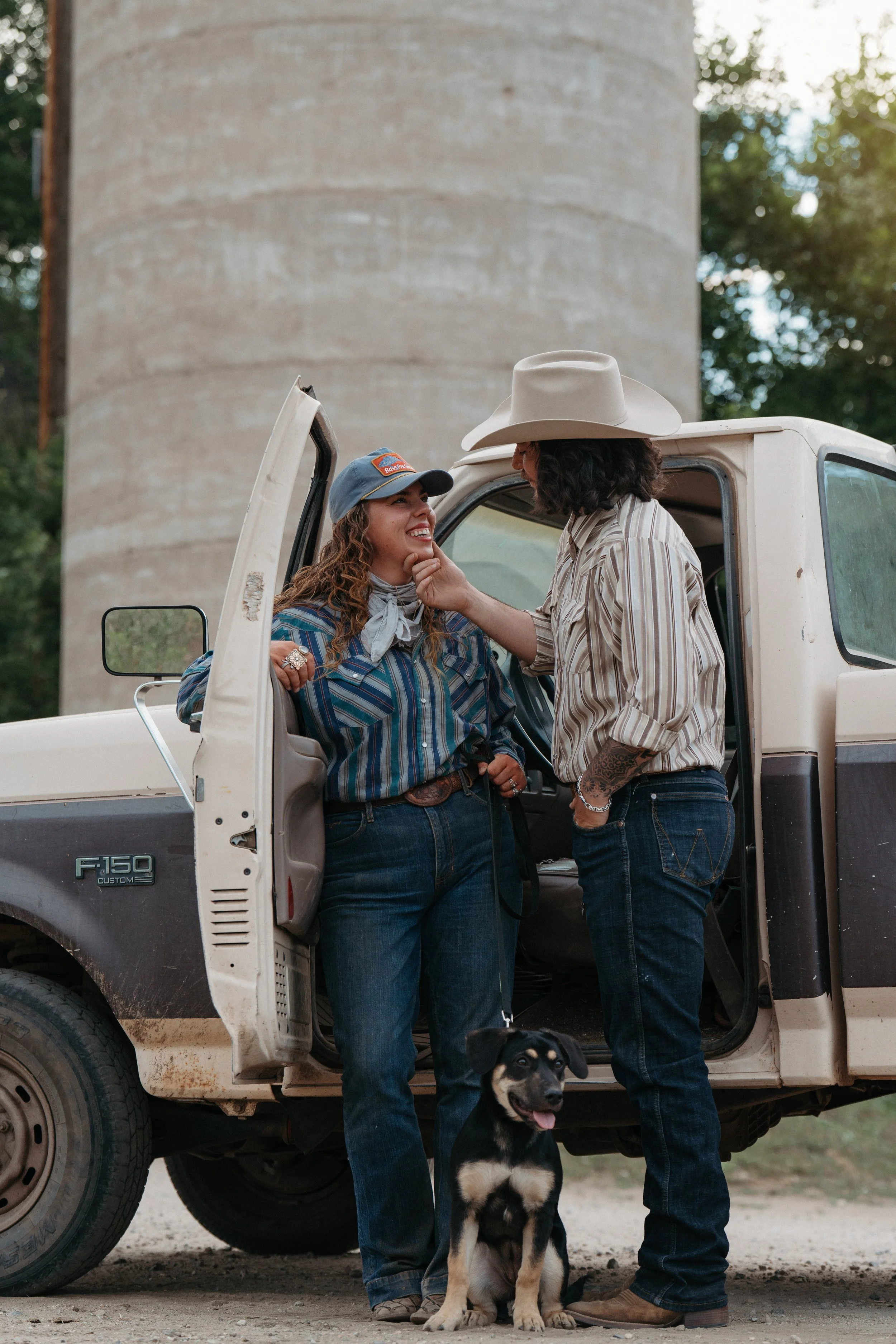 Two women standing beside a white pickup truck, talking and smiling. One woman is wearing a baseball cap and a striped shirt, and the other woman is wearing a cowboy hat and a striped shirt. A black and tan puppy is sitting on the ground in front of them. The background shows a large concrete silo and trees.
