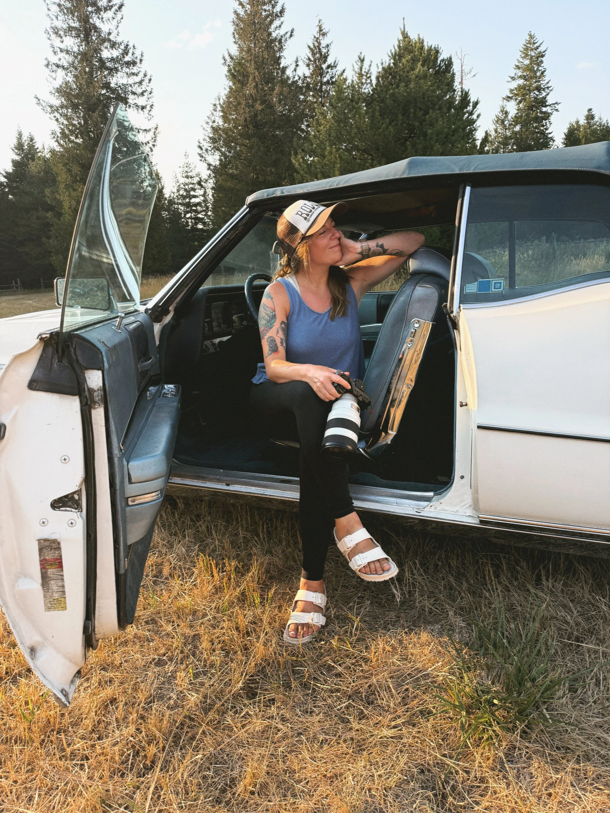 Young woman with tattoos, wearing a cap and blue tank top, leaning on an old white car with the door open in a grassy field with trees in the background, holding a large camera with a telephoto lens.