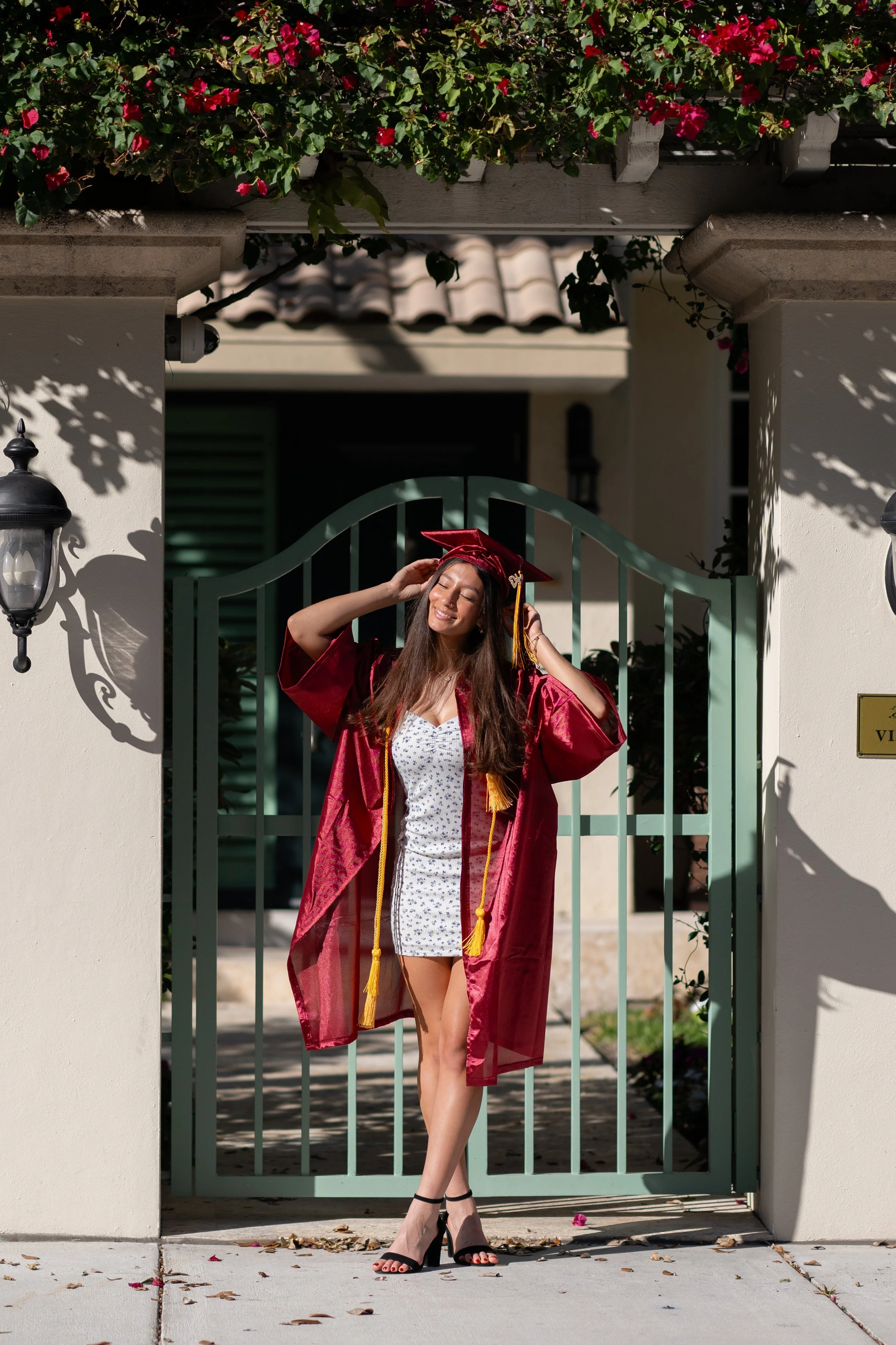 A young woman in a white dress with a floral pattern, wearing a red graduation gown and cap, stands happily at a gate, adjusting her cap in front of a white house with a tiled roof and pink flowers on a trellis above.