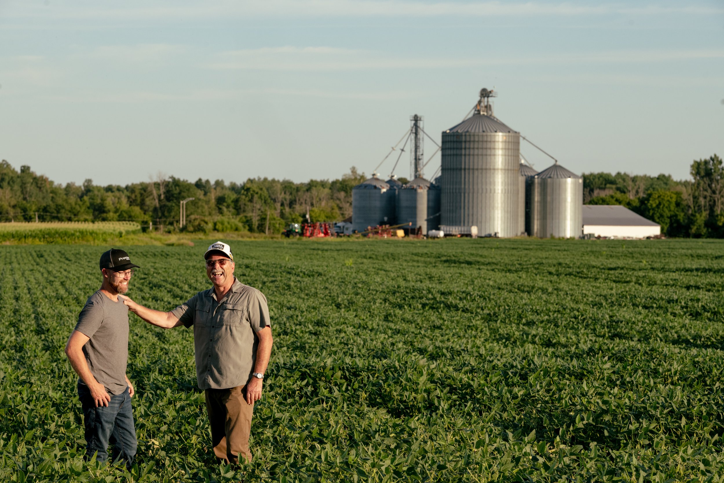 Two North Dundas farmers standing in a green farm field with silos and trees in the background, smiling and talking.