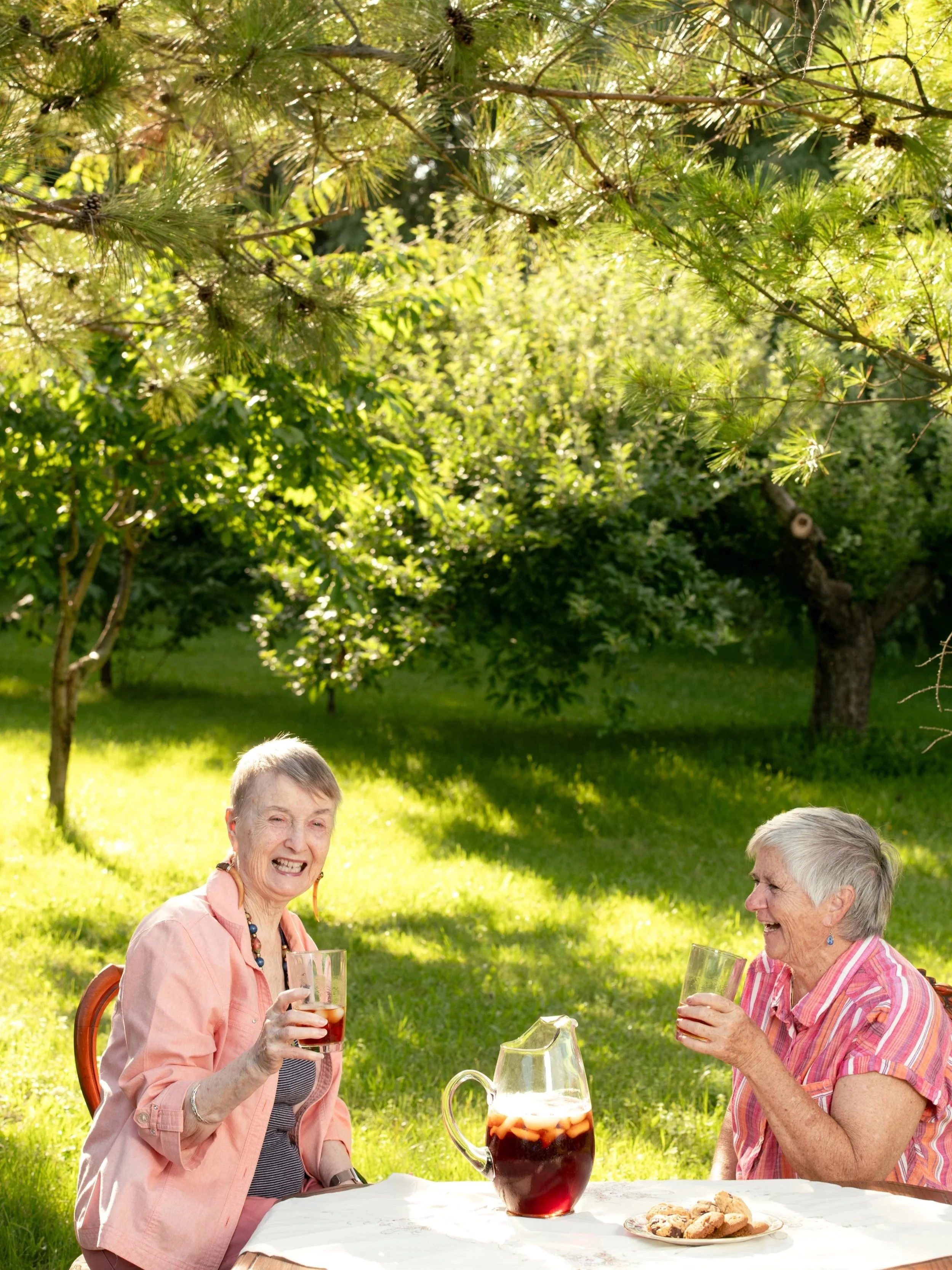 Theresa Bergeron with a friend, sitting at an outdoor table, smiling and laughing, in a garden setting.