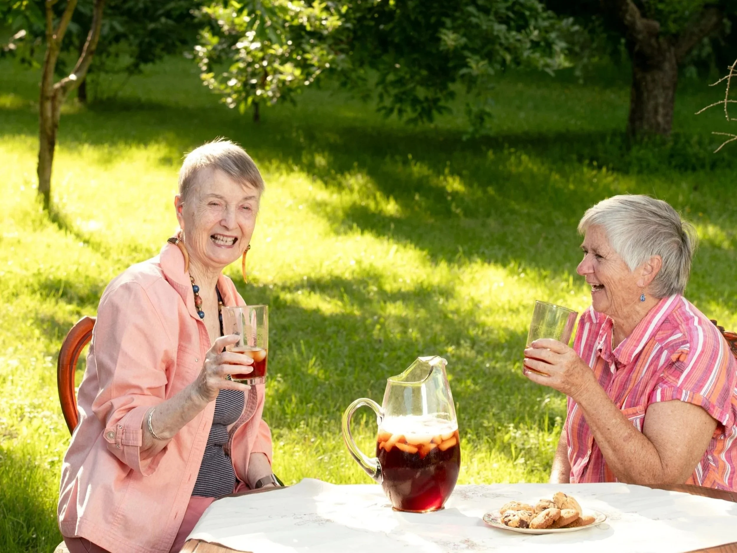 Theresa Bergeron with a friend, sitting at an outdoor table, smiling and laughing, in a garden setting.