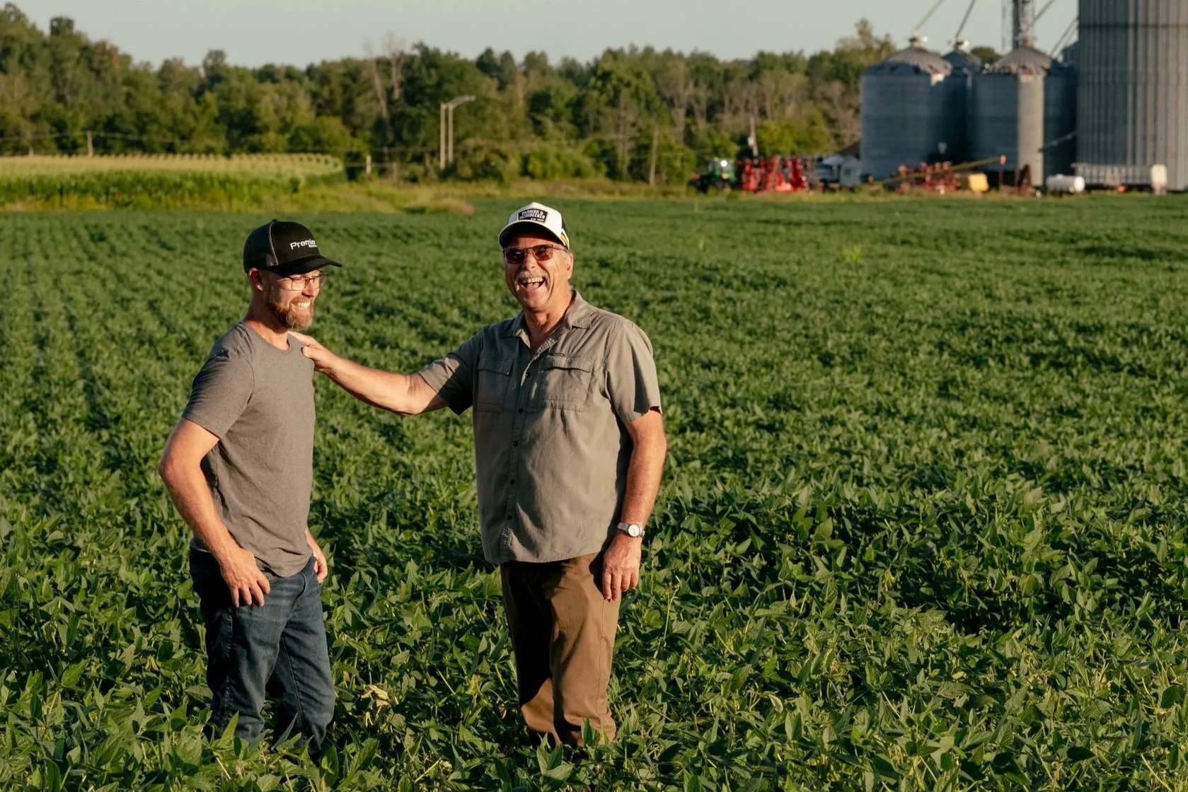 Two North Dundas farmers standing in a green farm field with silos and trees in the background, smiling and talking.
