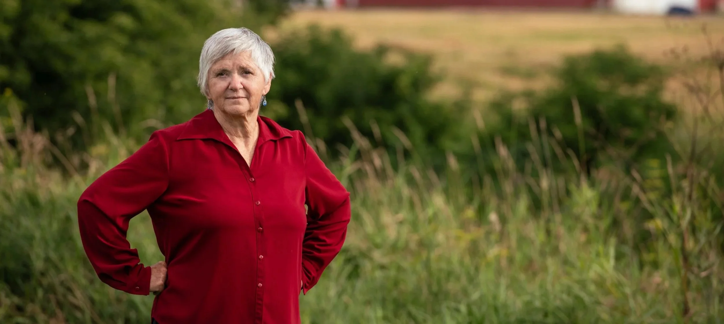 Theresa Bergeron, standing in a field with green foliage and a farm landscape in the background.