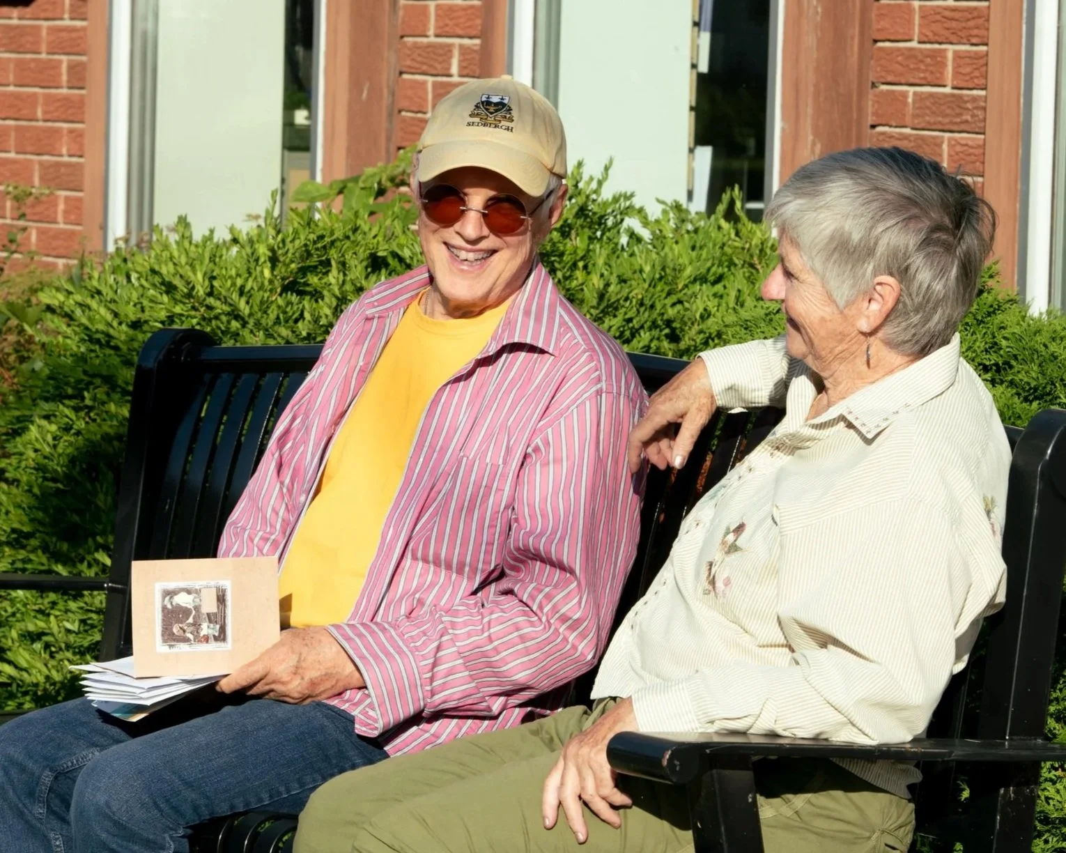 Two North Dundas seniors sitting on a bench outdoors, smiling and talking on a sunny day.