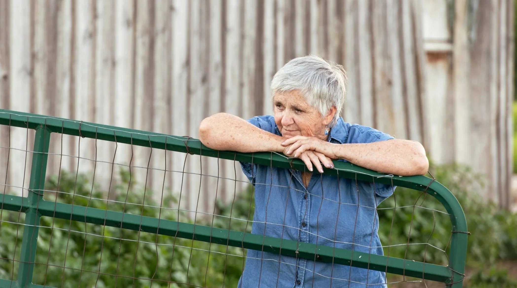 Theresa Bergeron, leaning on a green fence, resting her arms and looking thoughtfully into the distance.