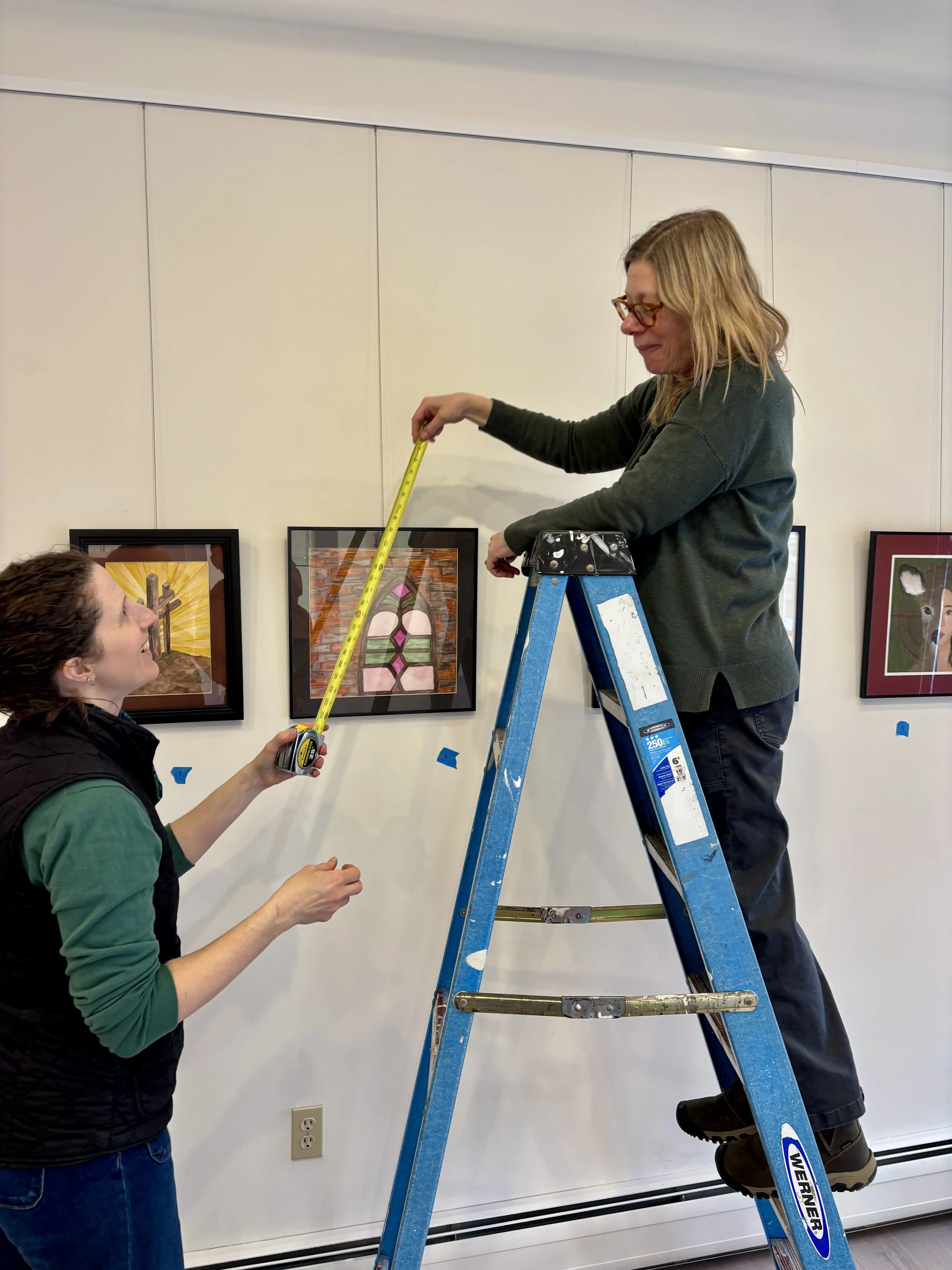 Two women are installing framed artwork on a white wall. One woman is standing on a blue ladder, holding a measuring tape, while the other woman is standing on the ground, assisting with the measurements.