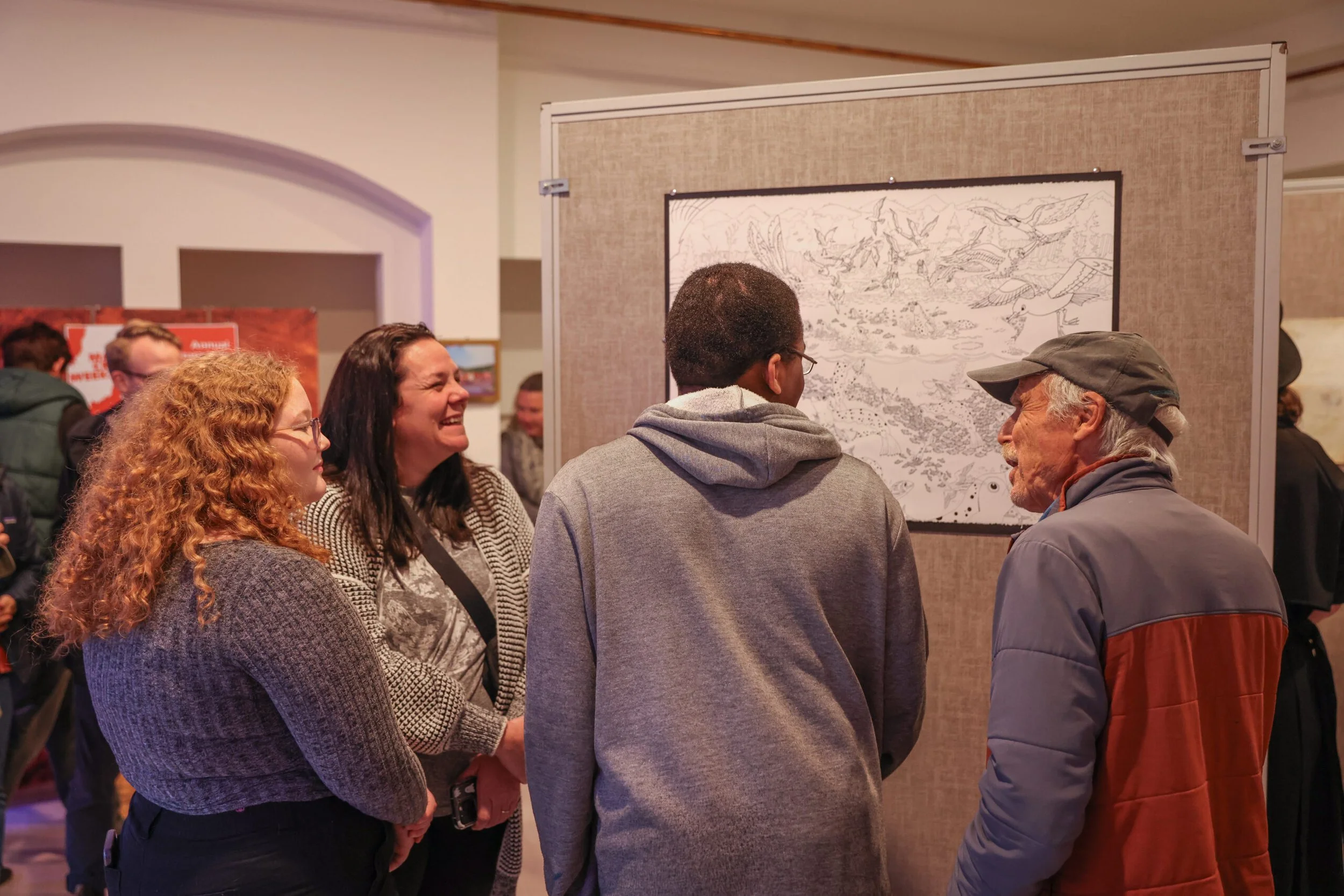 A group of five people, including three women and two men, is standing and smiling around a framed artwork in an art gallery.