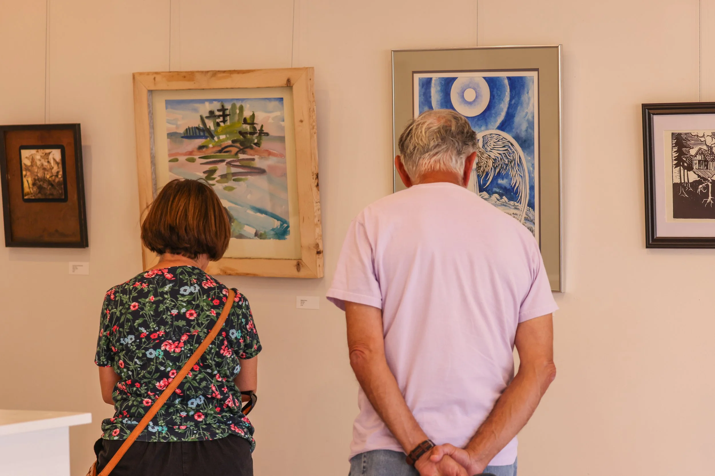 Two women and one man are looking at artworks in an art gallery. The woman on the left is wearing a black top with a floral pattern and has a tan purse strap over her shoulder. The man in the middle has gray hair and is wearing a pink shirt, standing