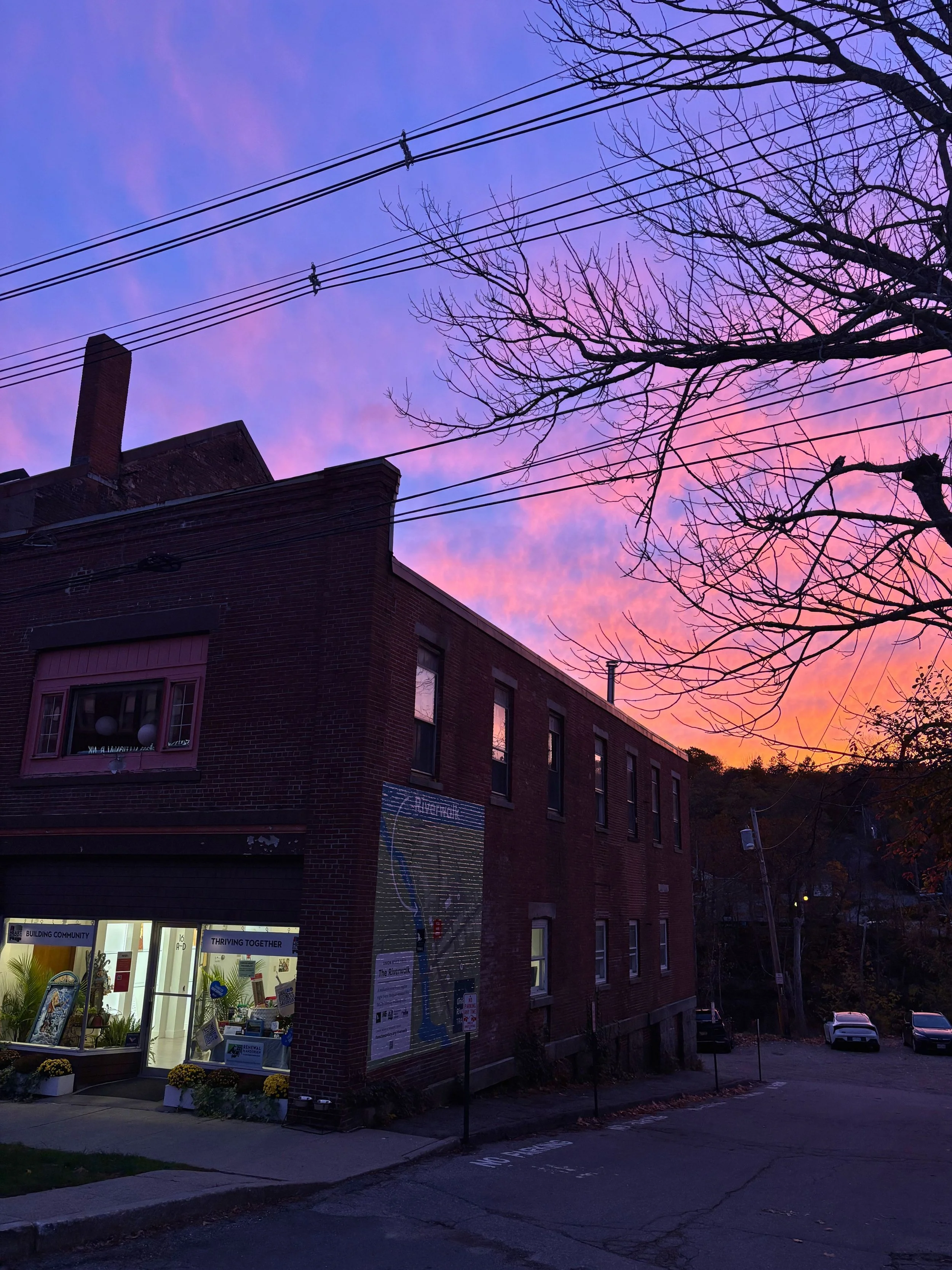 A brick building with large windows and a garage door on the ground floor, with a sign that says 'Building Community' and 'Thriving Together.' The sky is colorful with shades of purple, pink, and blue, and there are leafless trees in front.