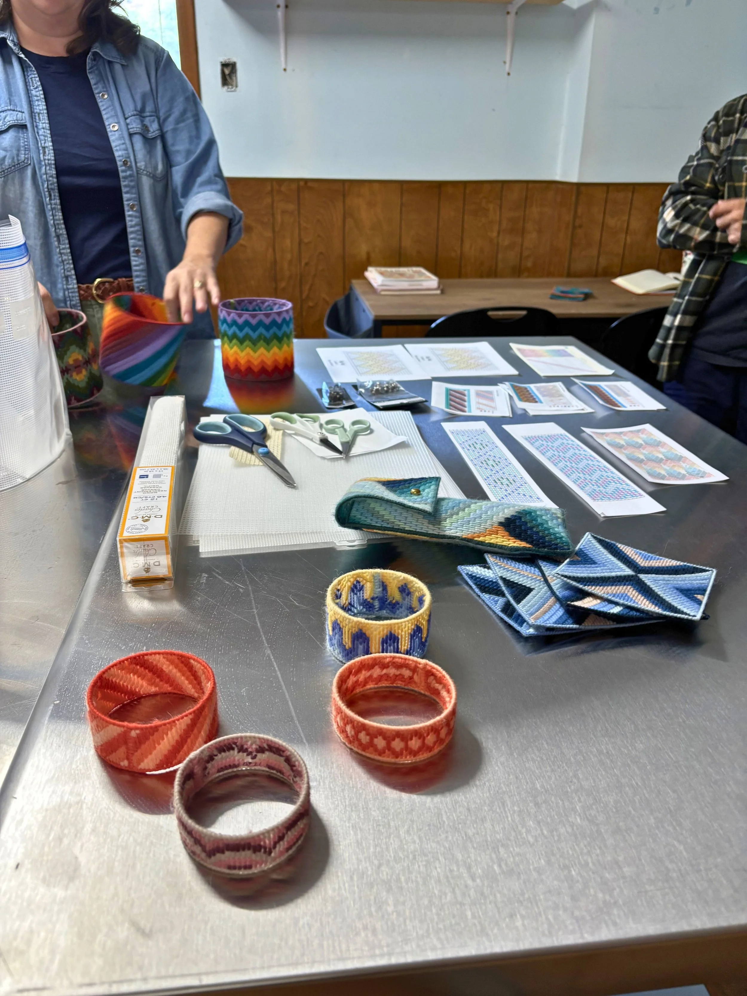 Colorful woven bracelets and fabric swatches laid out on a metal table, with papers, scissors, and quilting tools nearby.