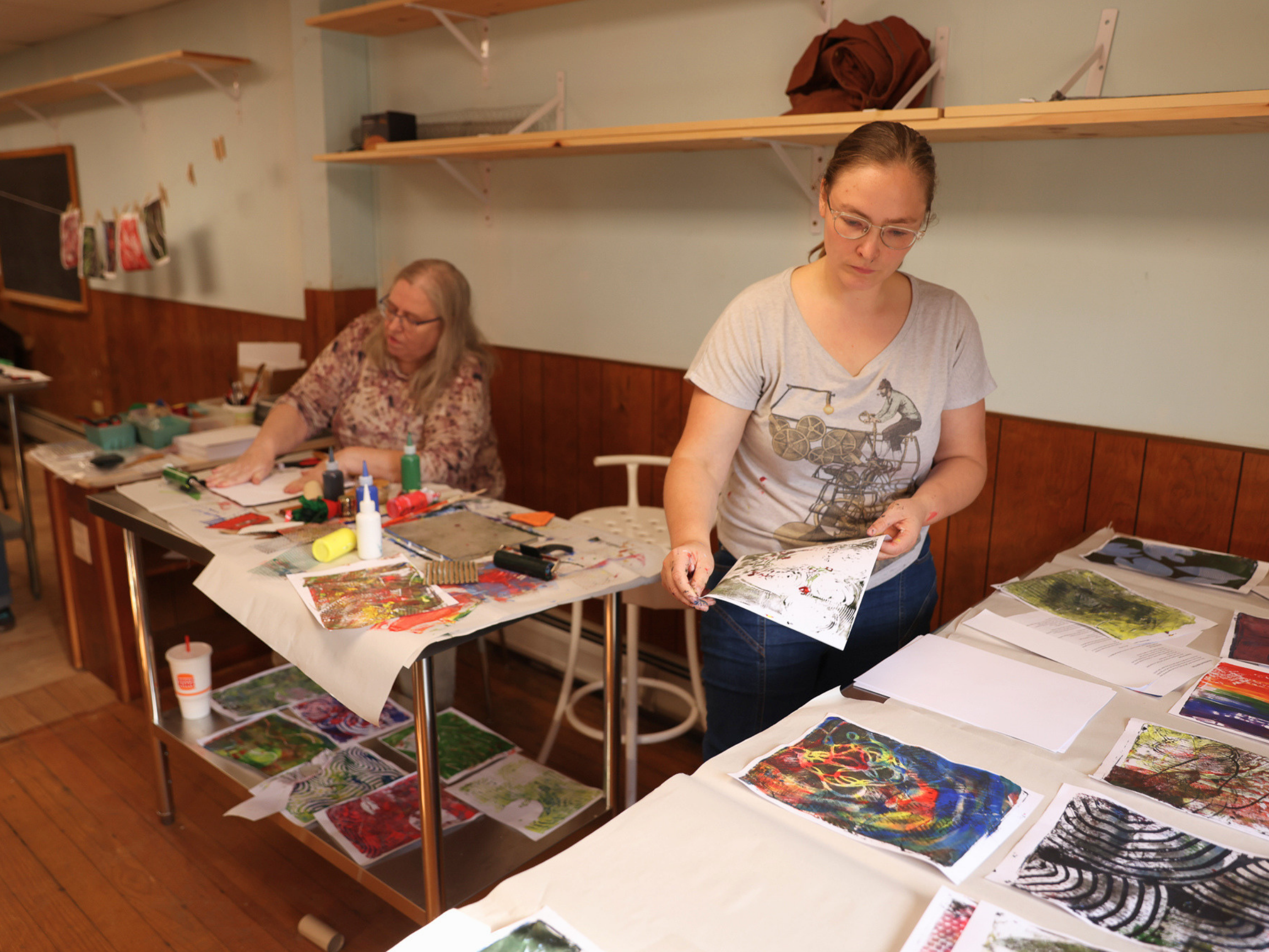 Two women working on art projects at a table covered with colorful artwork, paint bottles, and craft supplies in a room with wooden shelves and a chalkboard.