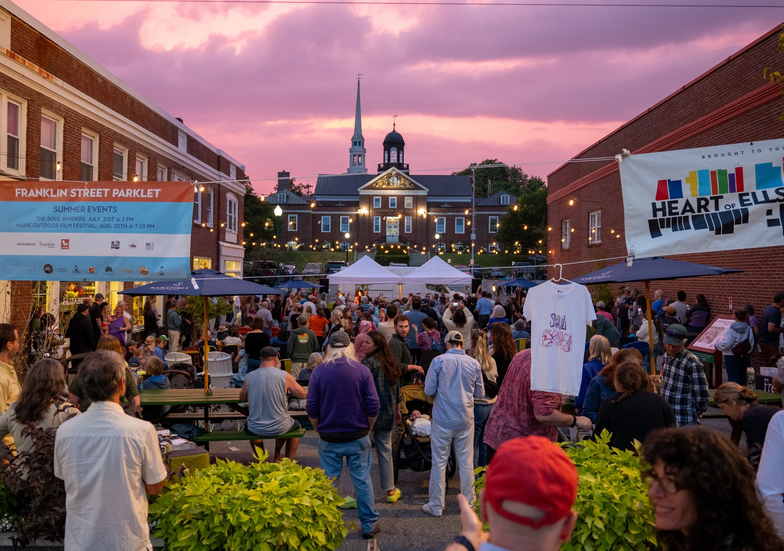 A lively outdoor community event during sunset with numerous people gathered around tables and tents, string lights hanging above, and a city hall building with a steeple and clock in the background under a pink and purple sky.
