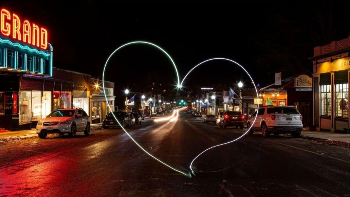 A night scene of a small town street illuminated by streetlights, with parked cars lining the road and storefronts on both sides. A heart shape is drawn with light in the foreground, adding a romantic atmosphere.