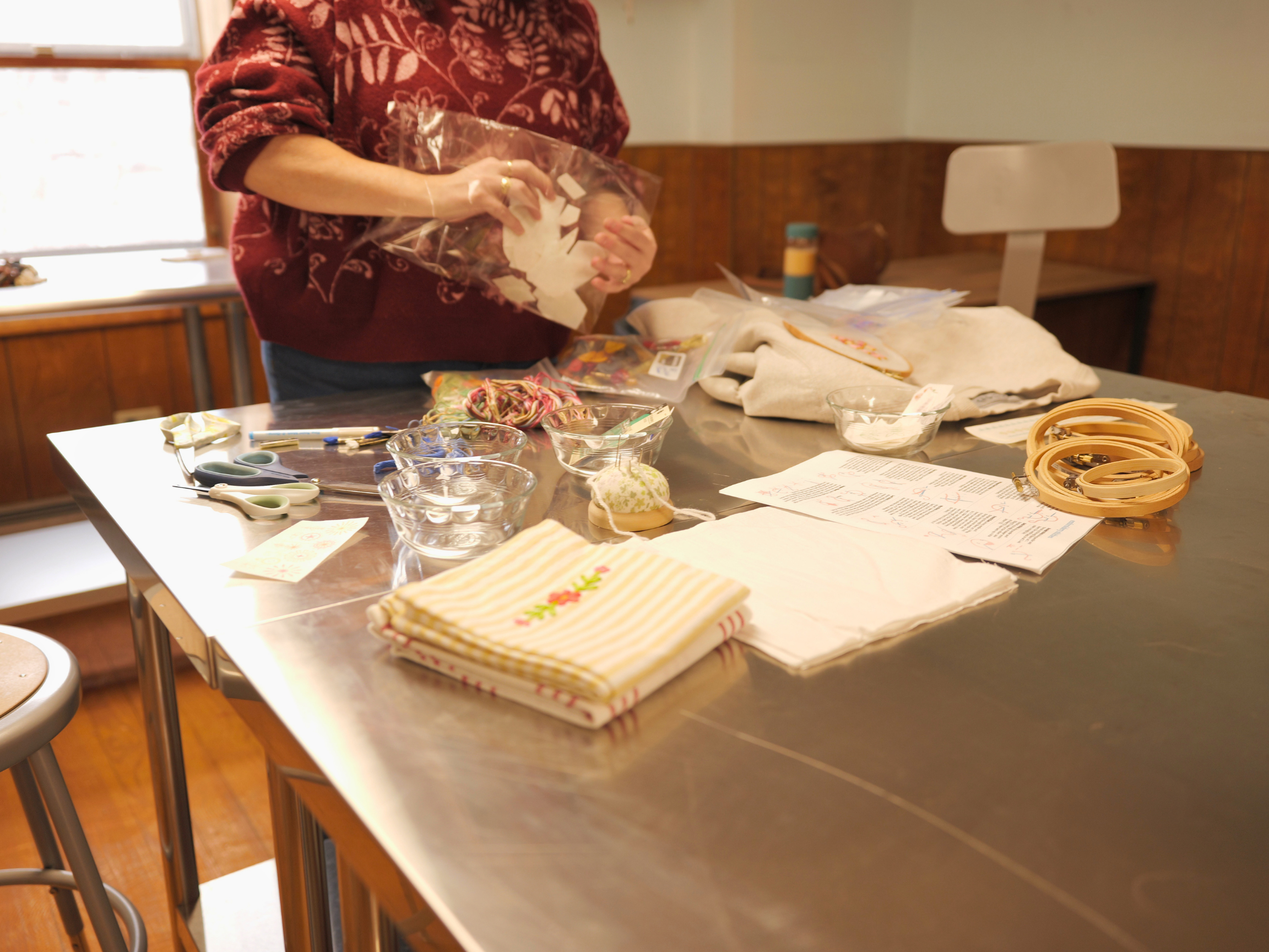 A person wearing a red floral sweater is standing at a table, wrapping a small gift or item in clear plastic. The table has various craft supplies including scissors, fabric, ribbons, and decorative items. In the background, there is a wooden wainscot and a chair.