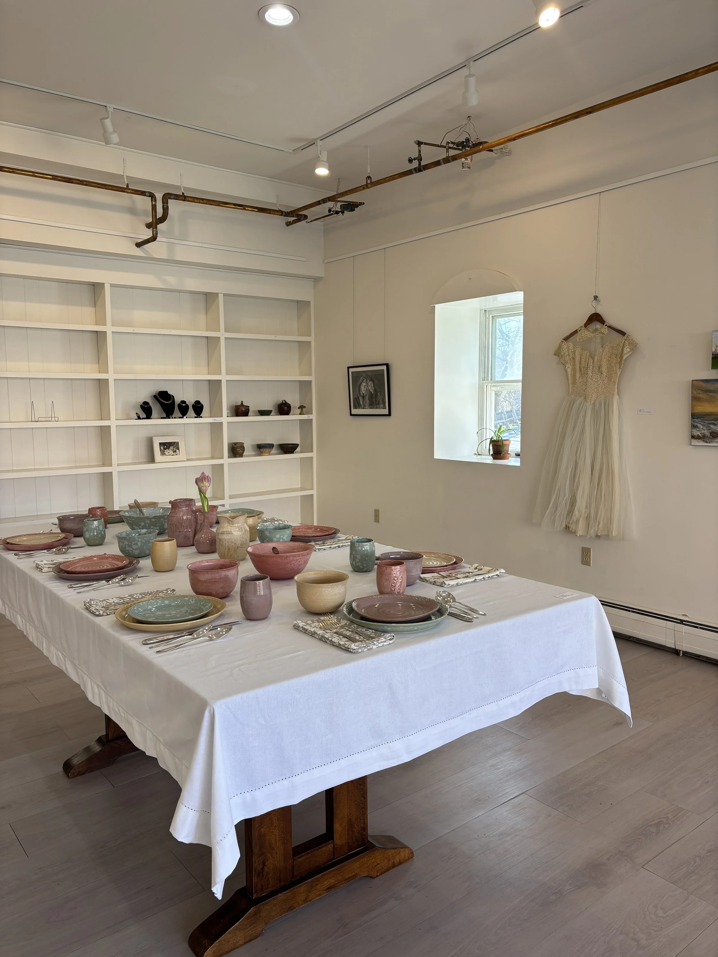 A dining table set with various colorful pottery, including bowls, plates, and cups, on a white tablecloth in a bright room with white walls, a shelf with decorative items, a framed picture, and a window with natural light.