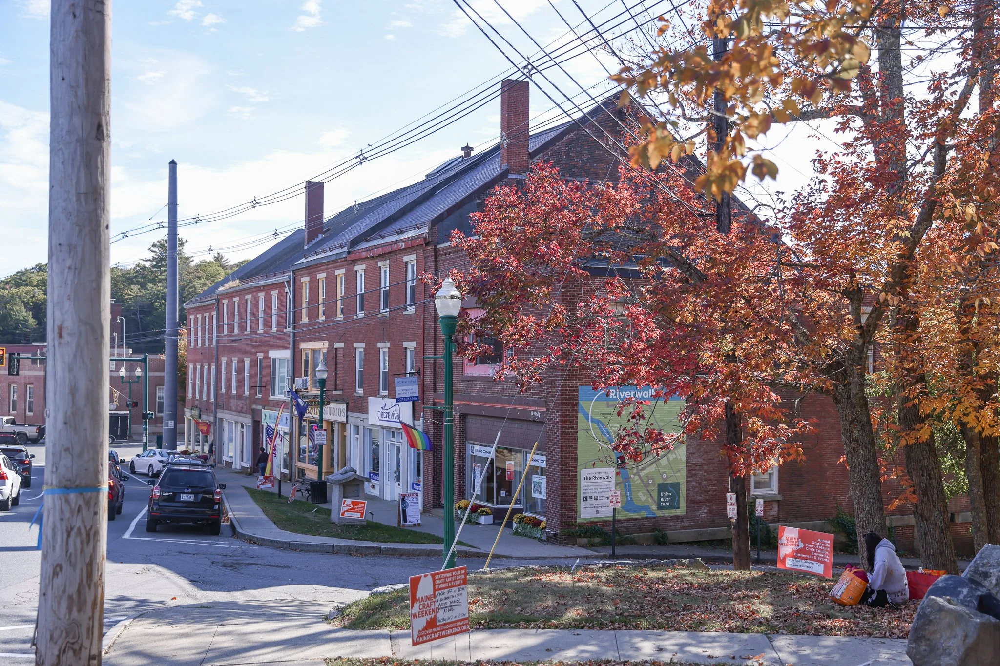 A street scene in a small town with a brick building, parking lot, trees with fall foliage, streetlights, utility poles, and signs for Maine Craft Weekend.