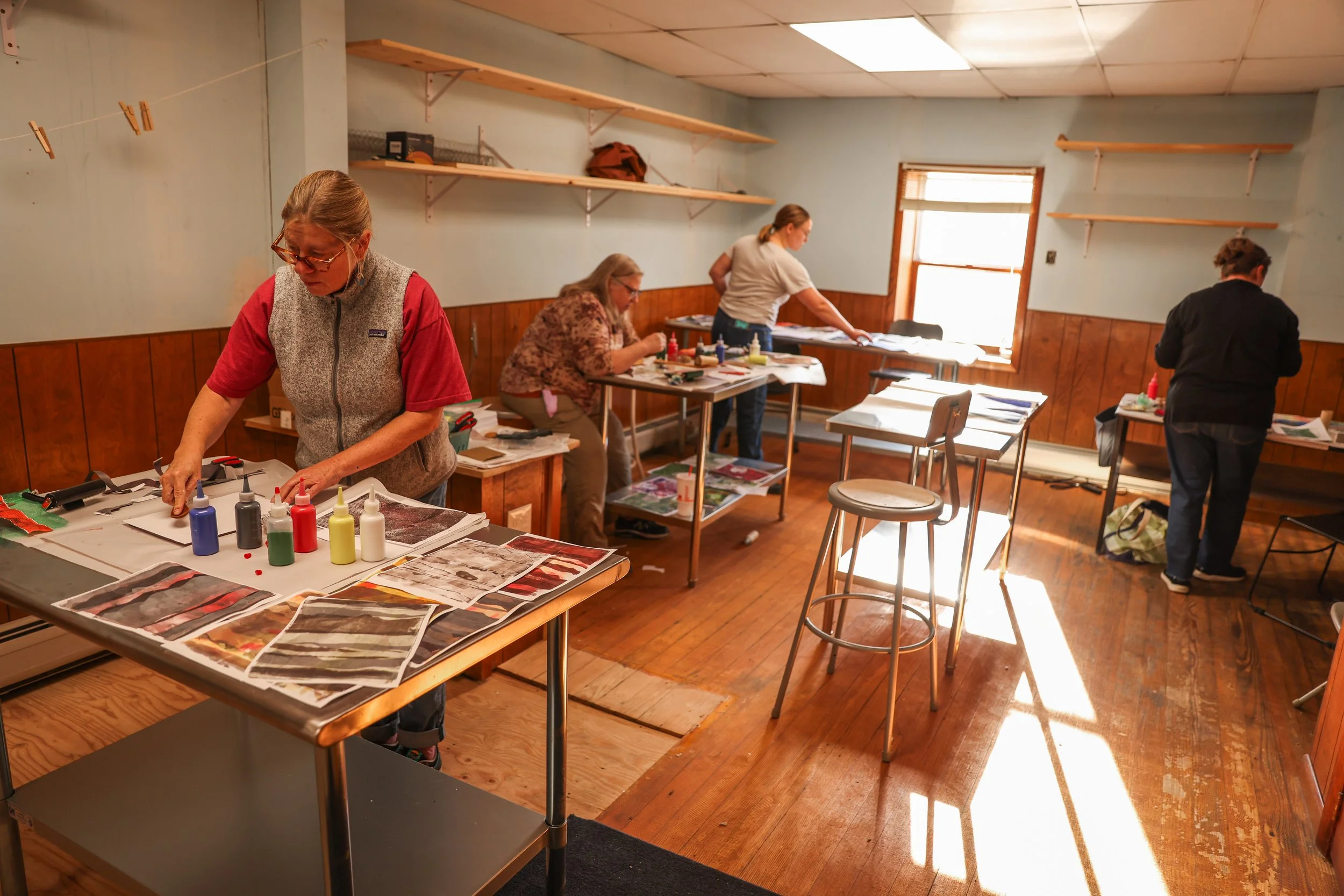 Women working on crafts in a brightly lit room with wood-paneled walls and shelves, supplies, and tables.