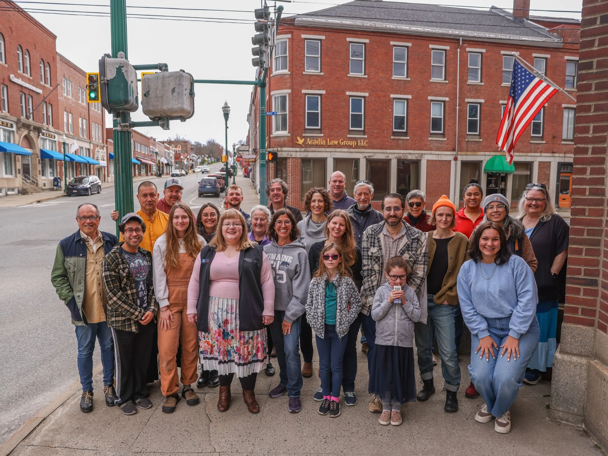 A diverse group of people, including men, women, and children, standing on a sidewalk in front of brick buildings on a city street. Some are smiling and looking at the camera, with a red brick building and American flag visible in the background.