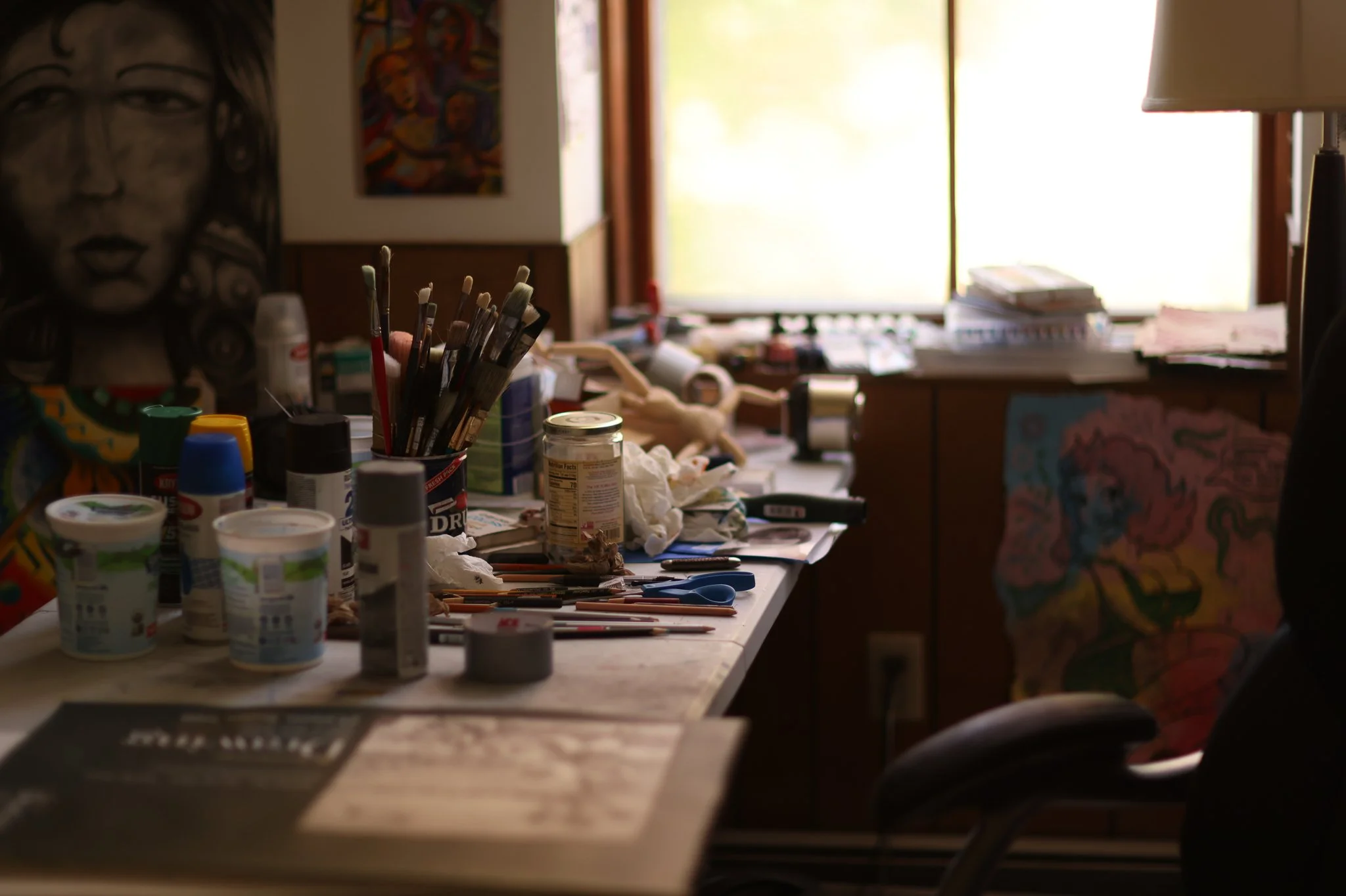 An artist's cluttered workspace with various paint bottles, brushes, and art supplies on a table, with a window letting in natural light and artwork hanging on the walls.